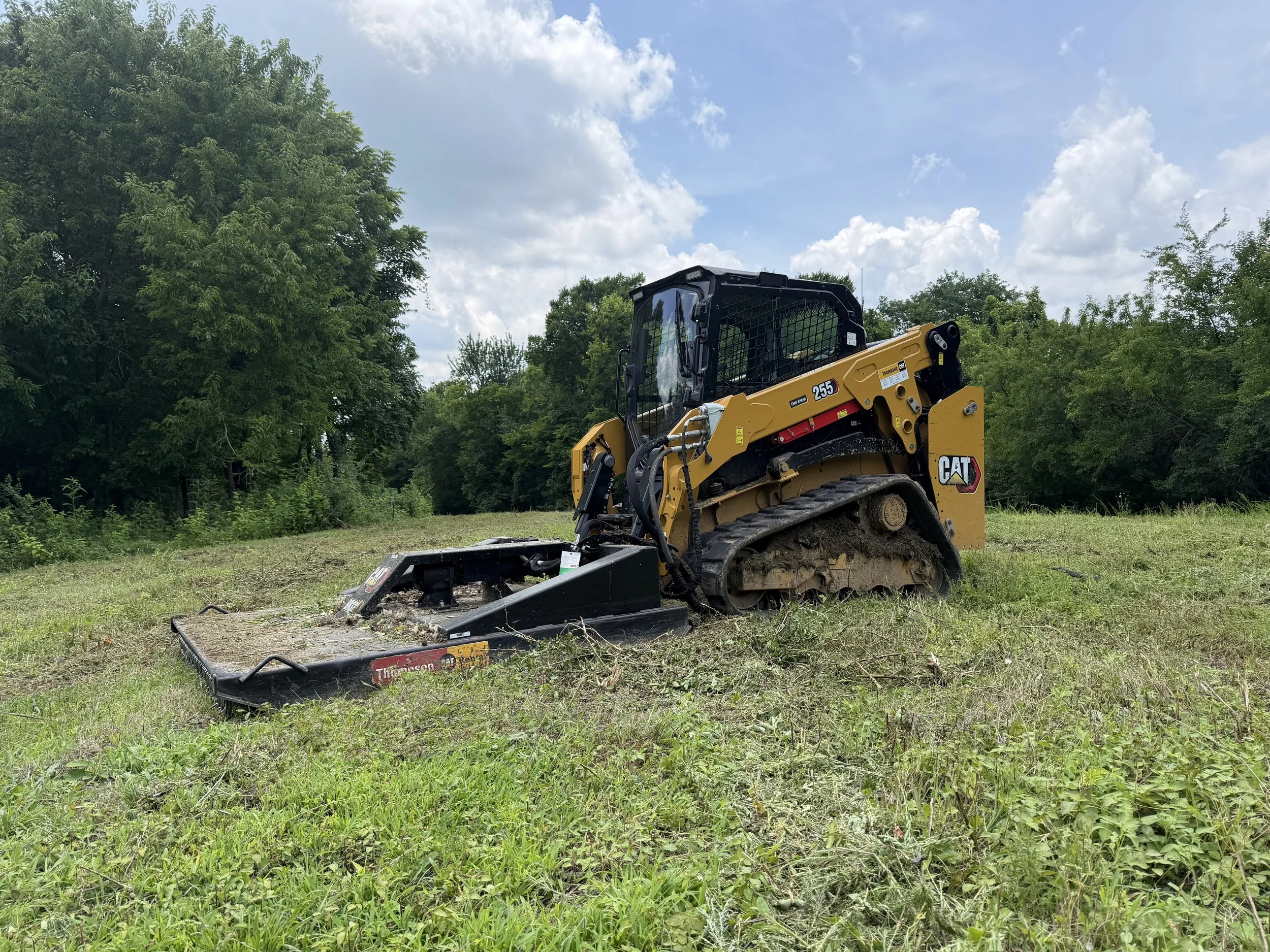 A yellow Caterpillar bulldozer working on a grassy area with trees and a partly cloudy sky in the background.