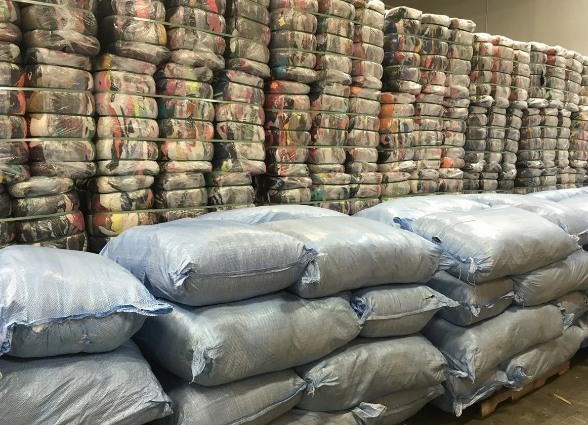 Large stacks of clear plastic bags filled with colorful fabric or clothing items, arranged on top of large gray bags in a storage room or warehouse.