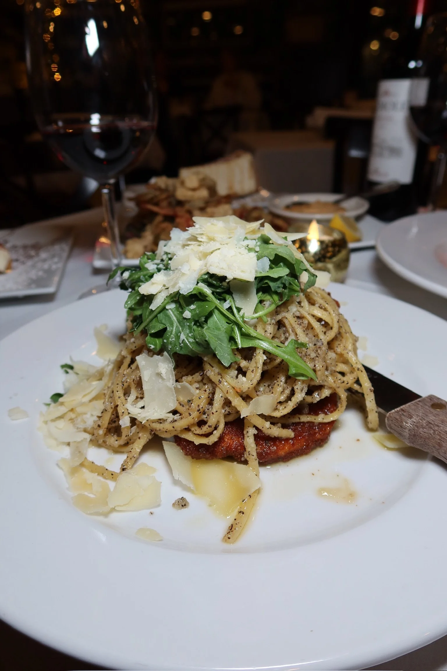 Plate of spaghetti pasta topped with arugula, shaved cheese, and black pepper. In a dimly lit restaurant setting with wine and bread in the background.