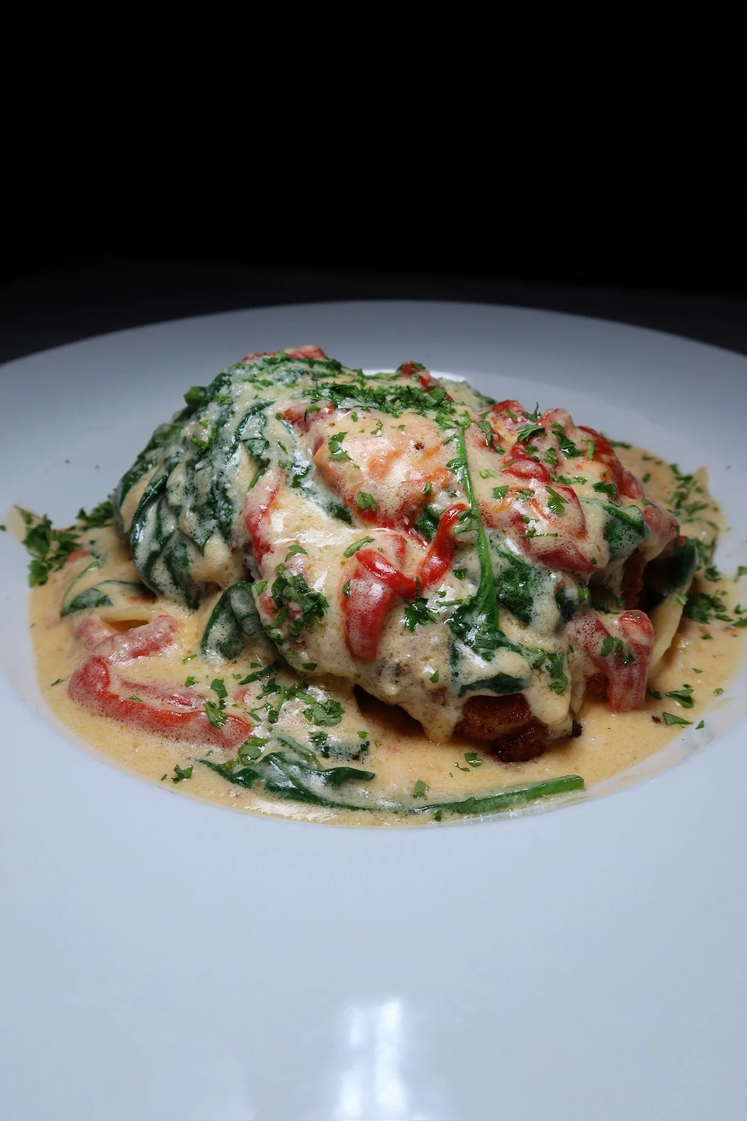 A close-up picture of a serving of chicken with a creamy sauce, chopped tomatoes, spinach, and parsley on a white plate.