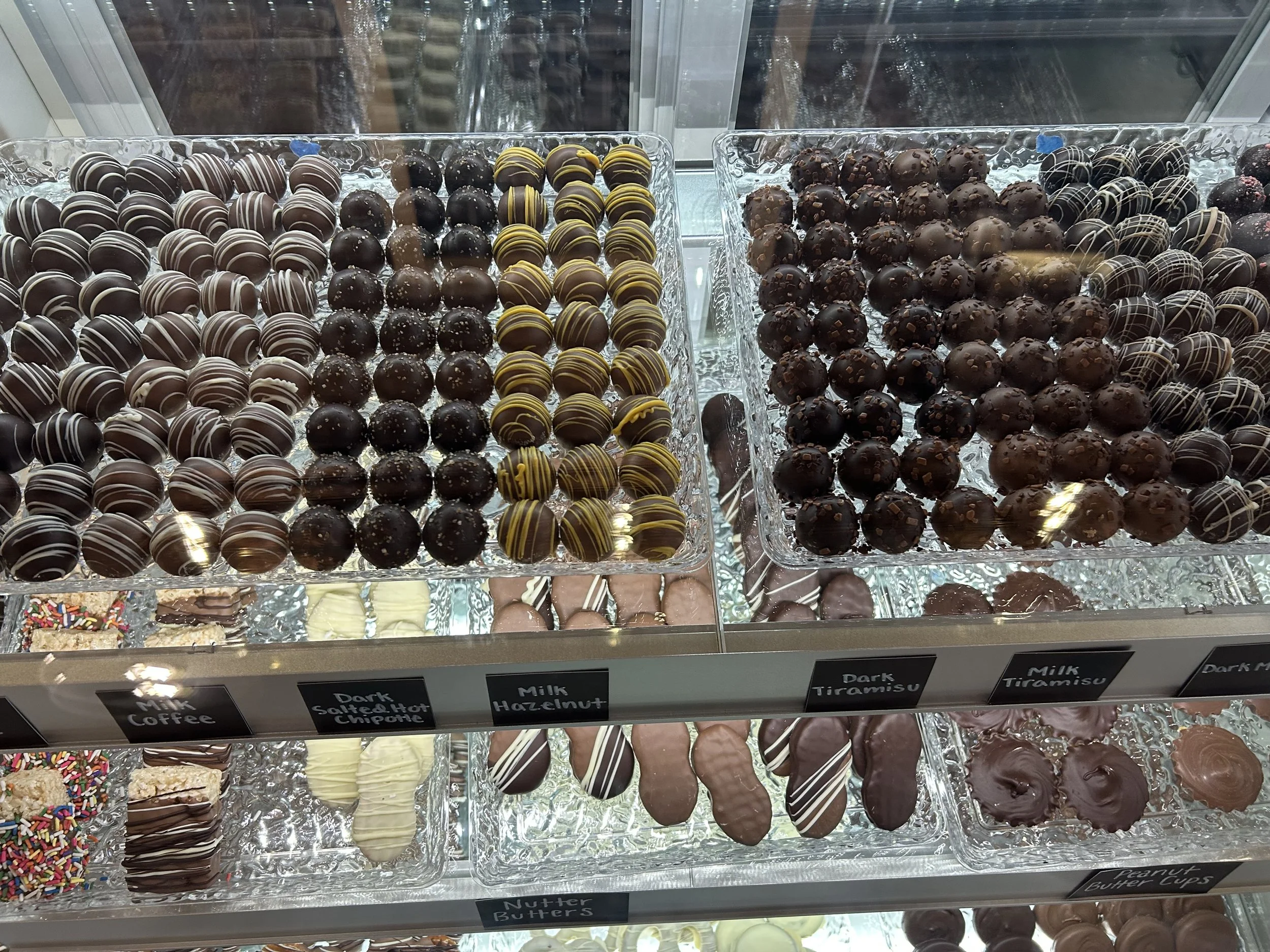 A display of assorted chocolates in a glass case. The chocolates are organized in rows, with labels at the bottom indicating flavors including Milk Coffee, Salted Hot Chipotle, Hazelnut, Dark Tiramisu, Milk Tiramisu, and Nutter Butters.