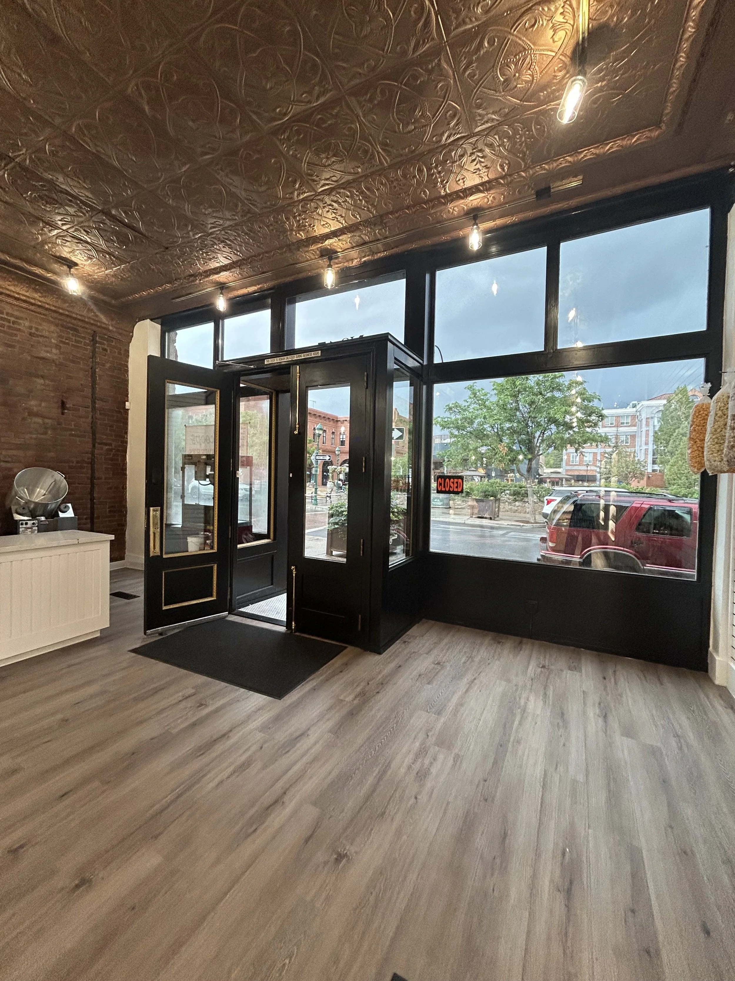 Inside view of a closed storefront with large glass windows and a glass door, a wooden floor, and a ceiling with decorative tin tiles and hanging light bulbs. Through the windows, a street scene with buildings, trees, and parked cars is visible.