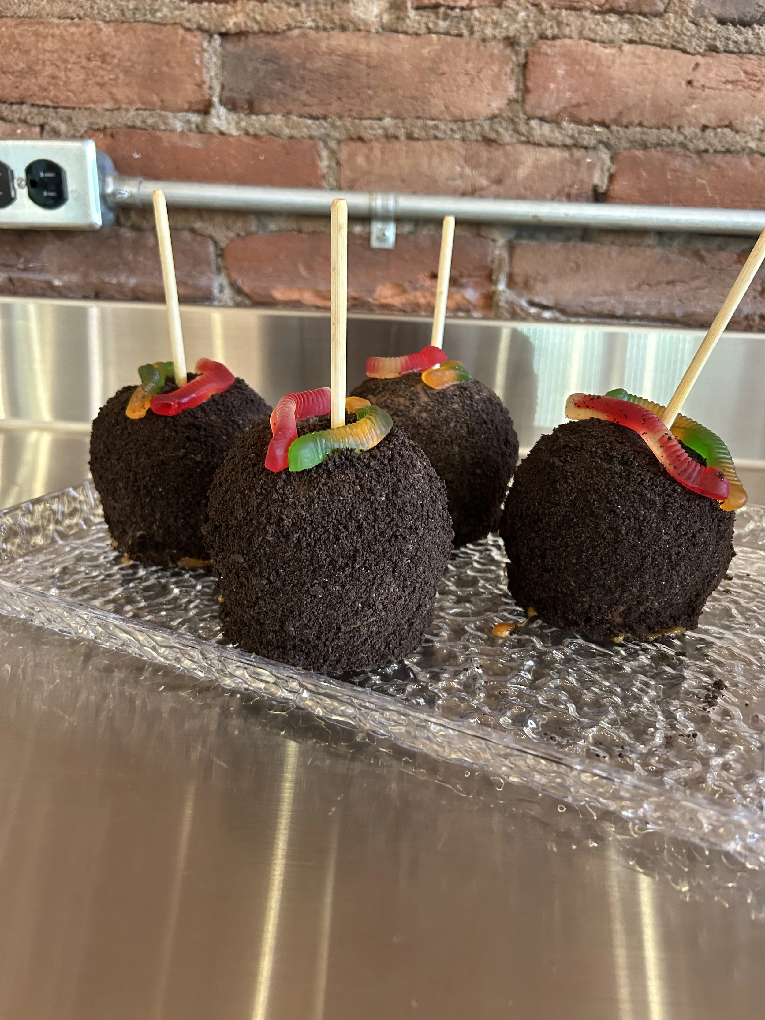 Four caramel apples coated in black crumbs, decorated with colorful gummy worms, and skewered with sticks, placed on a reflective silver surface with a brick wall background.