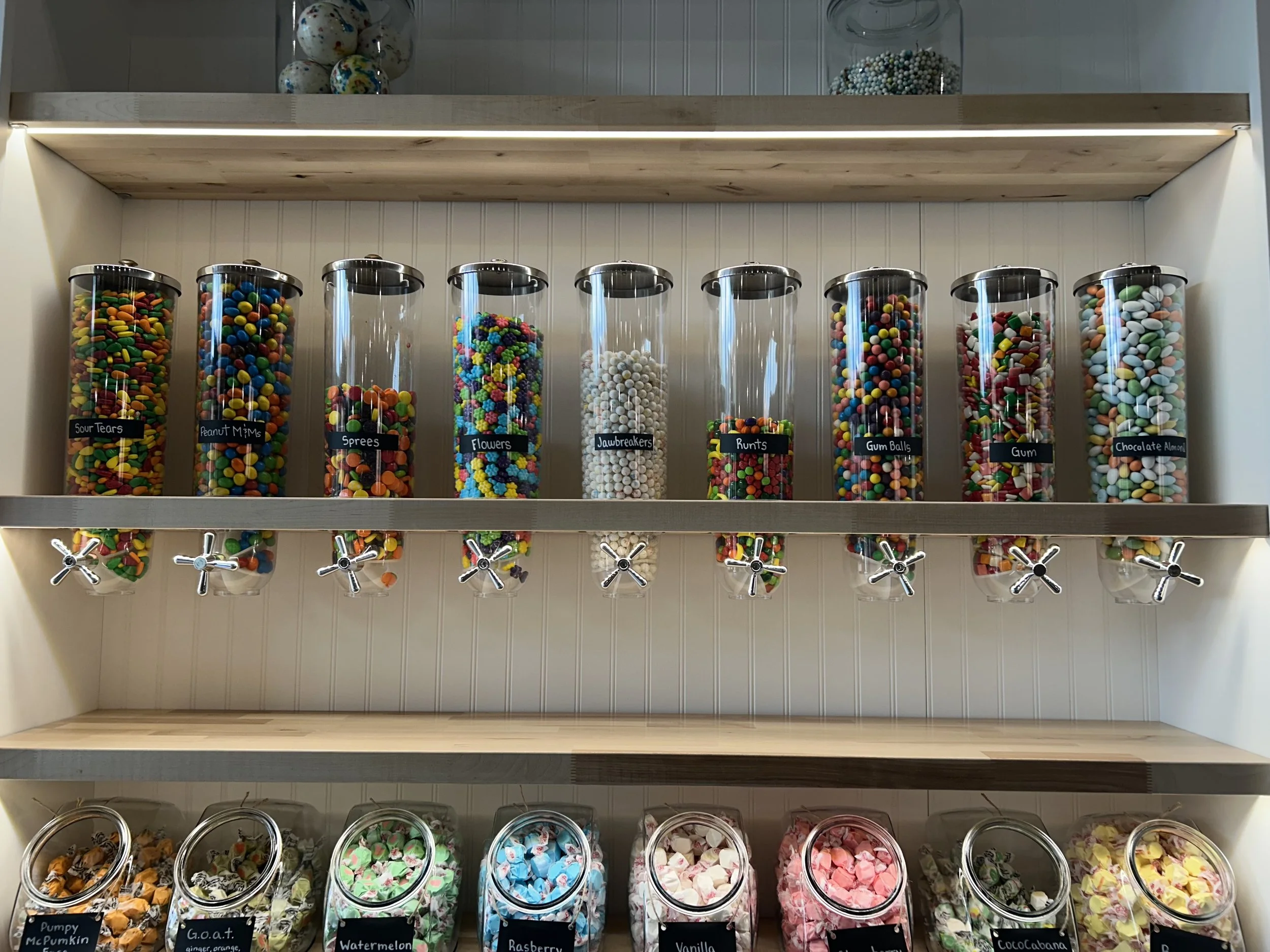 Shelf of large glass jars filled with colorful candies, each labeled with different candy names such as Sour Tears, Peanut M Ms, Sweets, Flowers, Jawbreakers, Runtz, Gum Balls, and Chocolate Almonds, with candies in various colors and shapes.