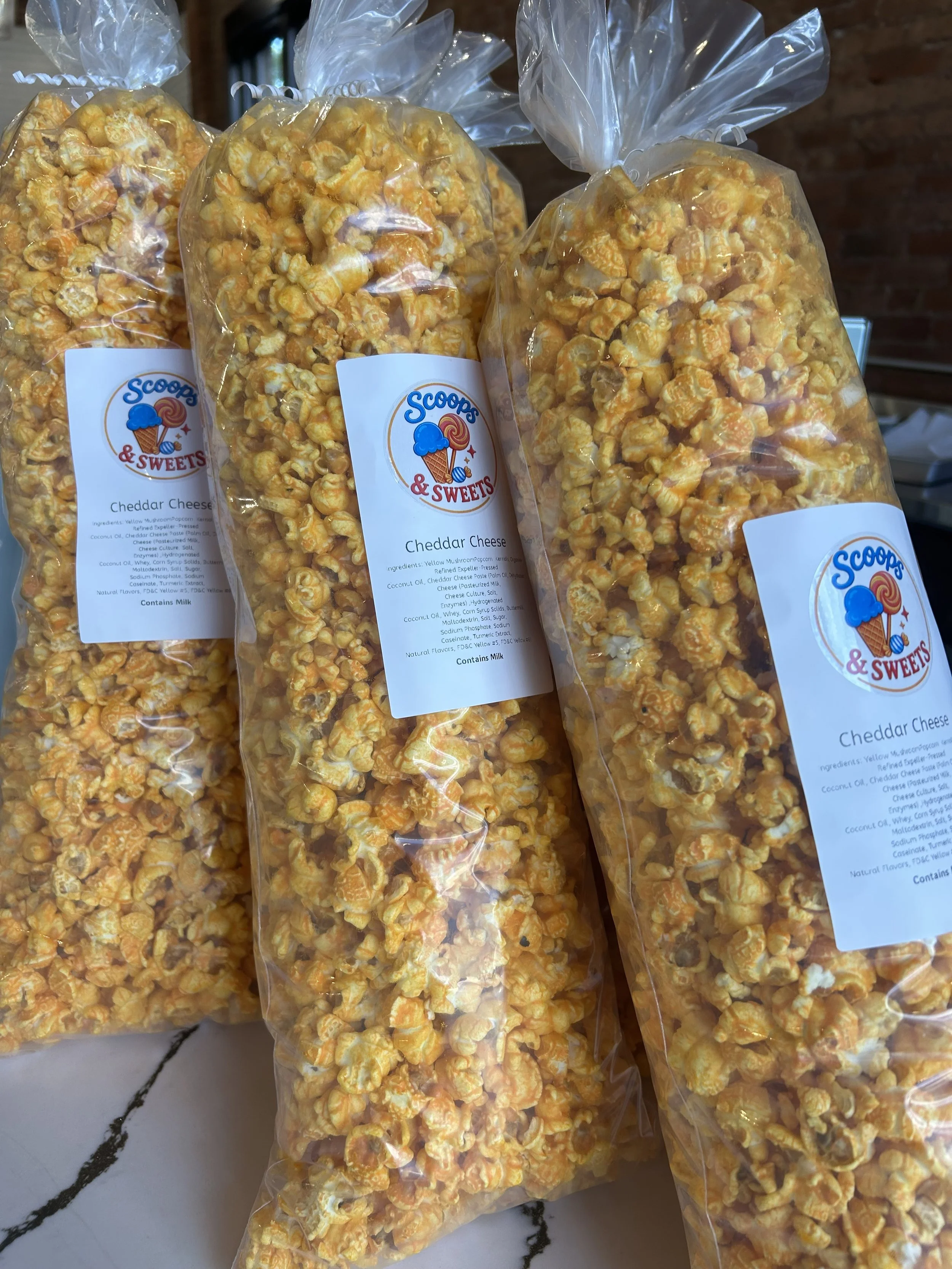 Several clear plastic bags filled with cheddar cheese popcorn, labeled 'Scoops & Sweets Cheddar Cheese', placed on a marble surface.