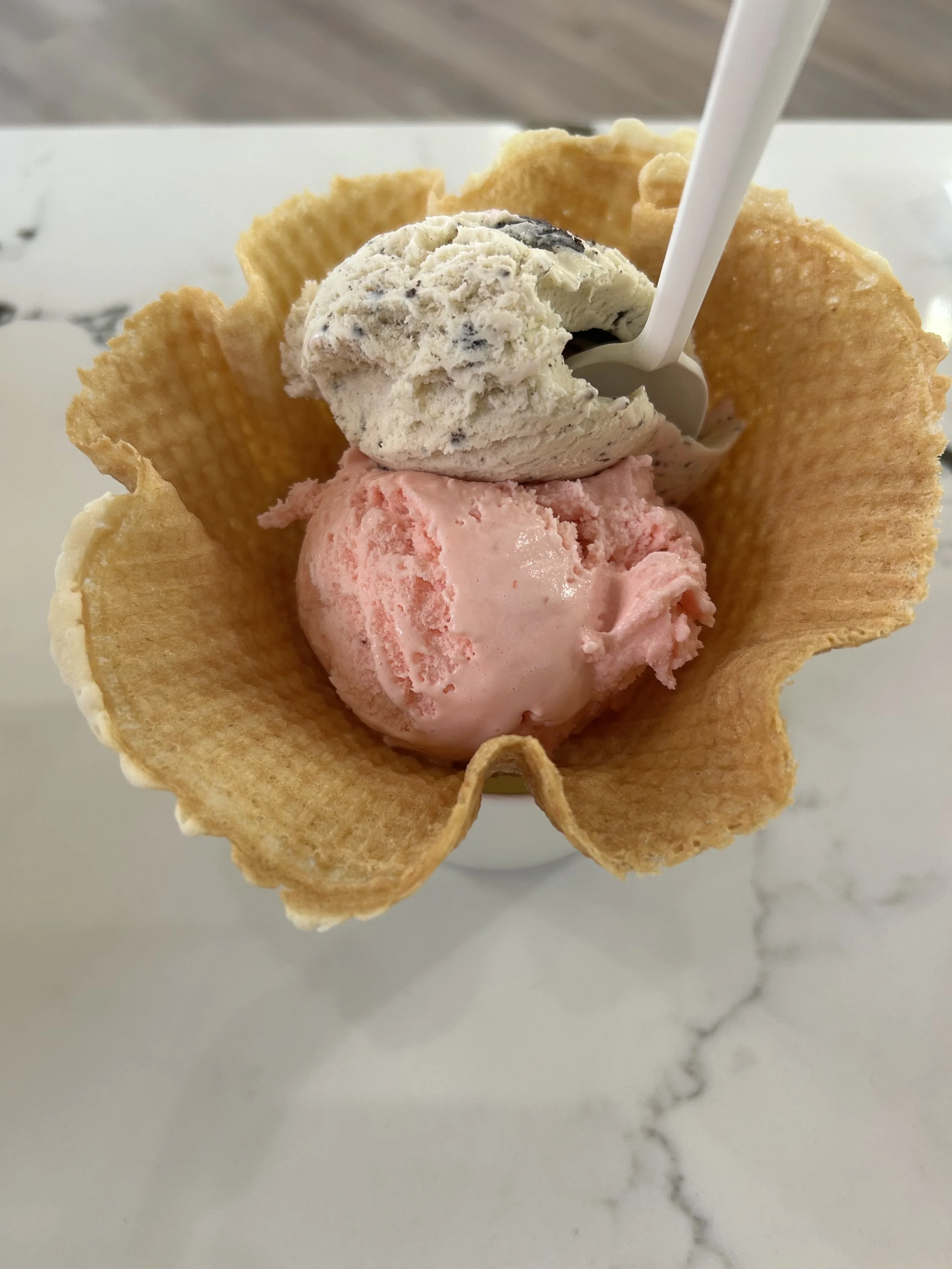 two scoops of ice cream in a waffle bowl with a white plastic spoon, on a white marble surface.