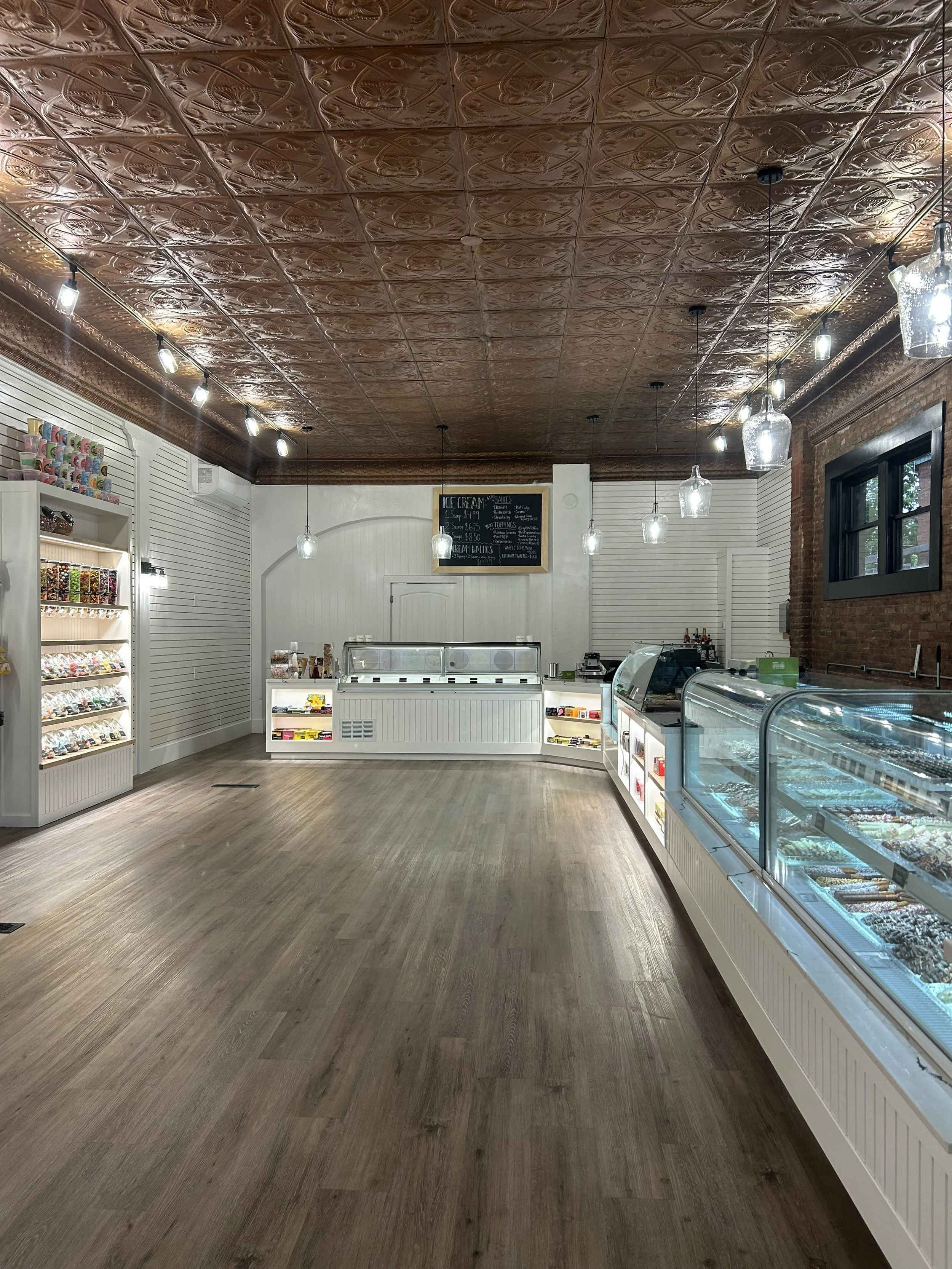 Interior of a candy or ice cream shop with a decorative embossed tin ceiling, string lights, brick and white walls, a chalkboard menu, and display cases filled with candies and ice cream.