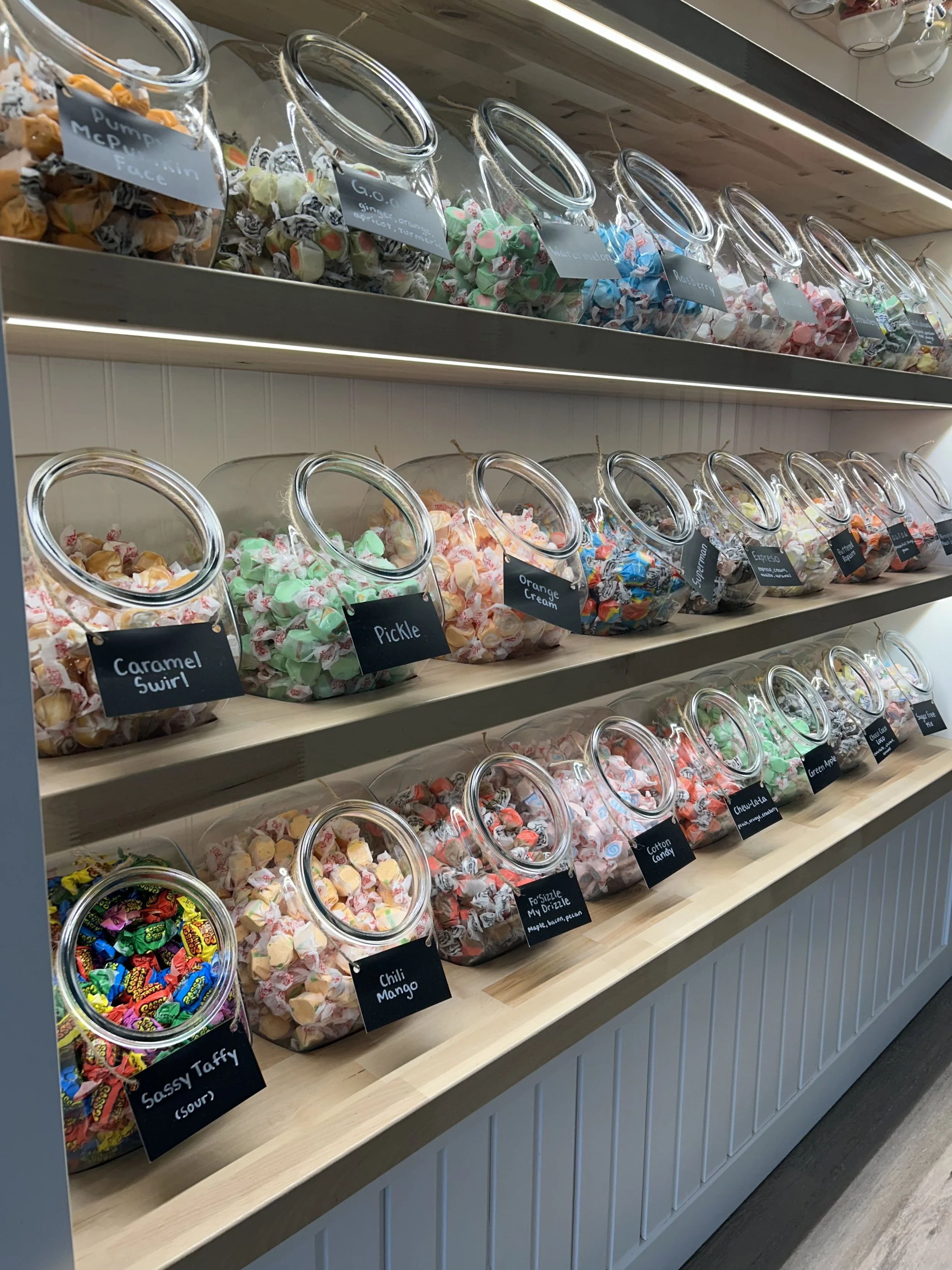 Display of assorted candies in glass jars labeled with their flavors, including caramel swirl, pickle, sassy taffy, orange cream, cotton candy, espresso, and chili mango, with a wooden shelf underneath.