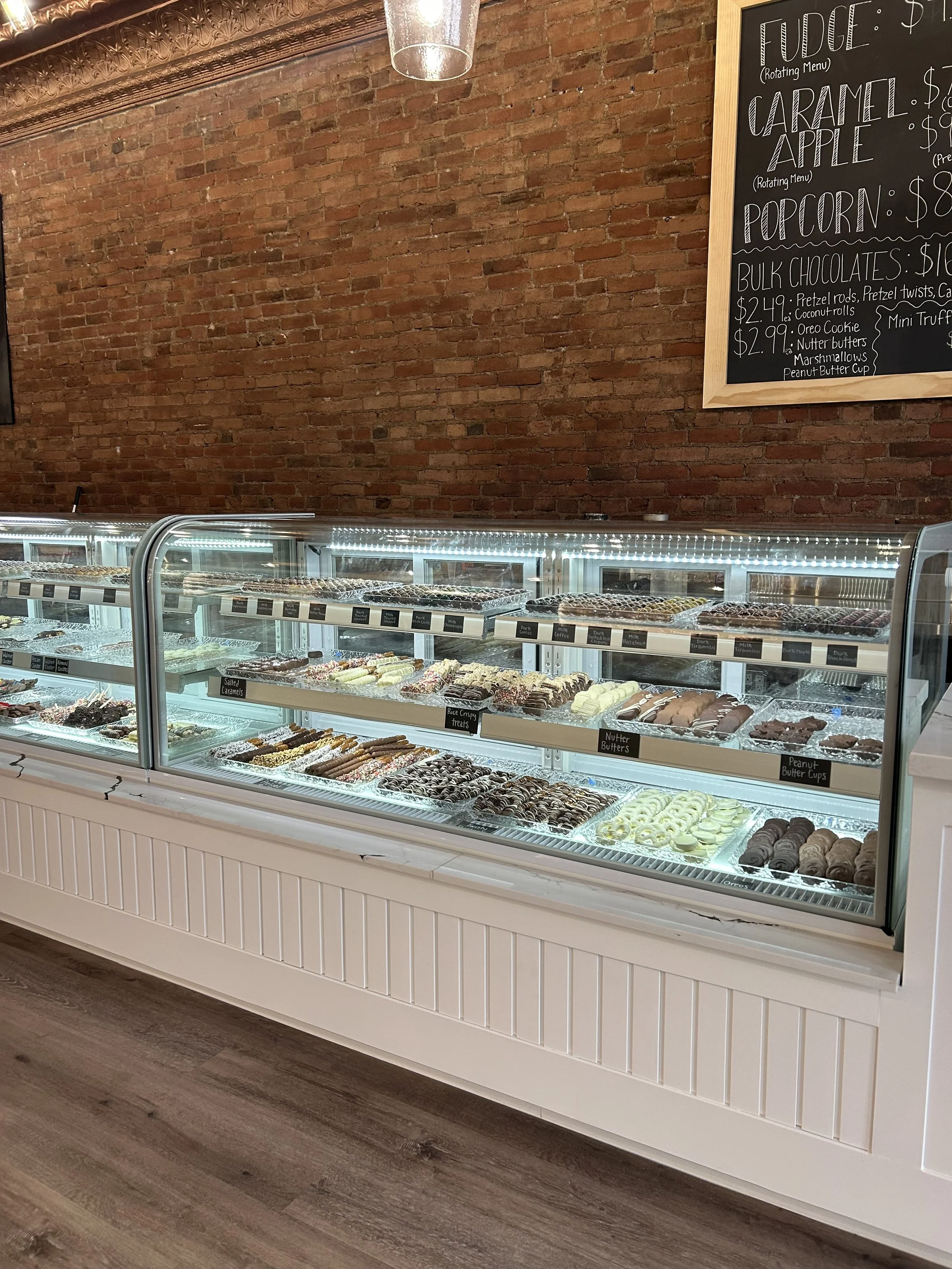 Display case filled with assorted chocolates and confections in a store with a brick wall background and blackboard menu.