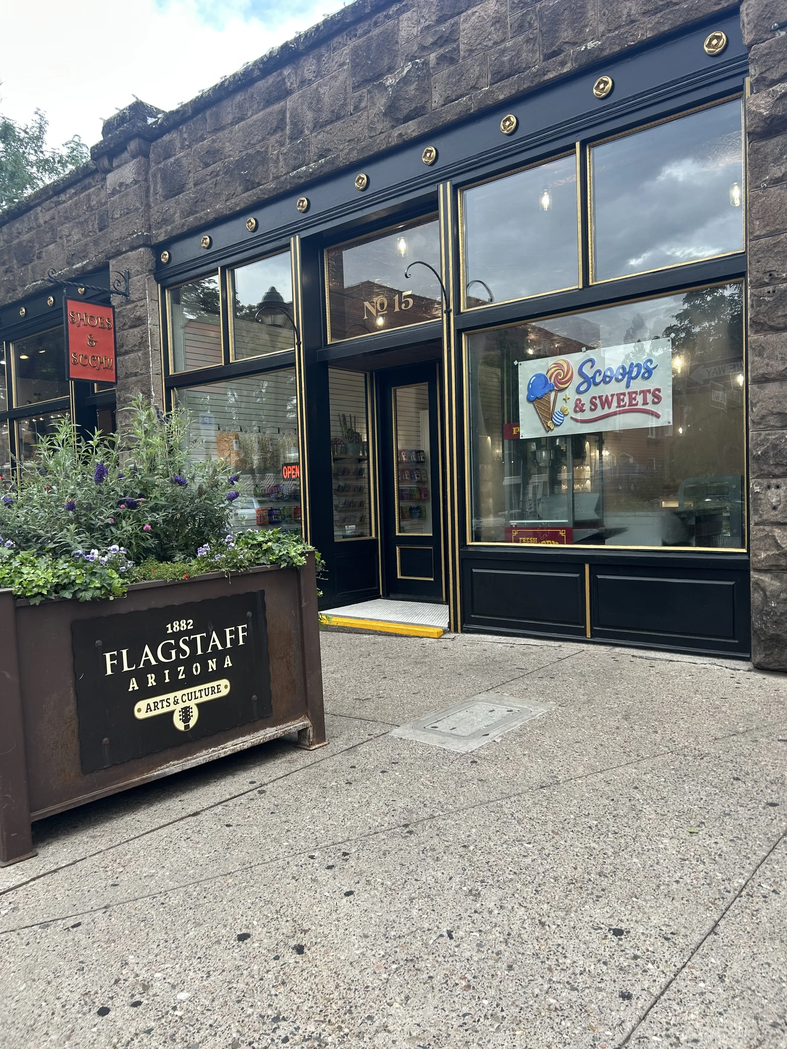 Exterior view of a storefront called Scoops & Sweets with large glass windows and a black and gold trim. In front, there is a flower planter with purple and green plants. The sidewalk is visible, and the sky is partly cloudy.