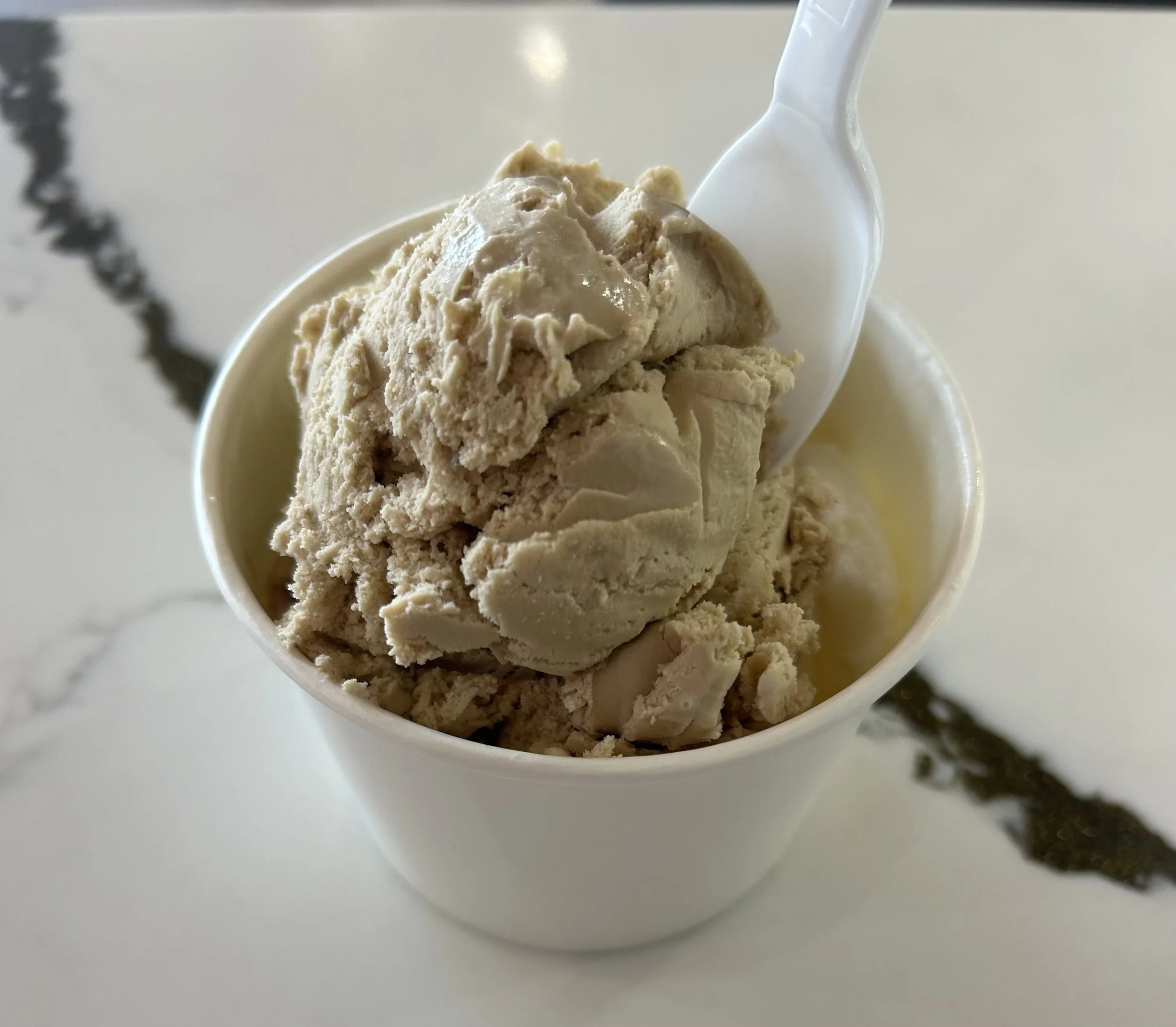 A cup of ice cream with a light brown color topped with a white spoon and small bits of the same ice cream.