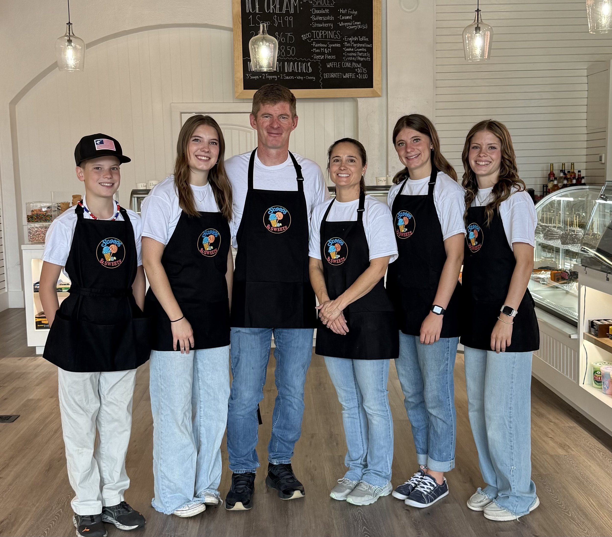 the owners and their 4 kids standing behind a counter in an ice cream shop, all wearing matching black aprons with a colorful ice cream logo, smiling for the camera.