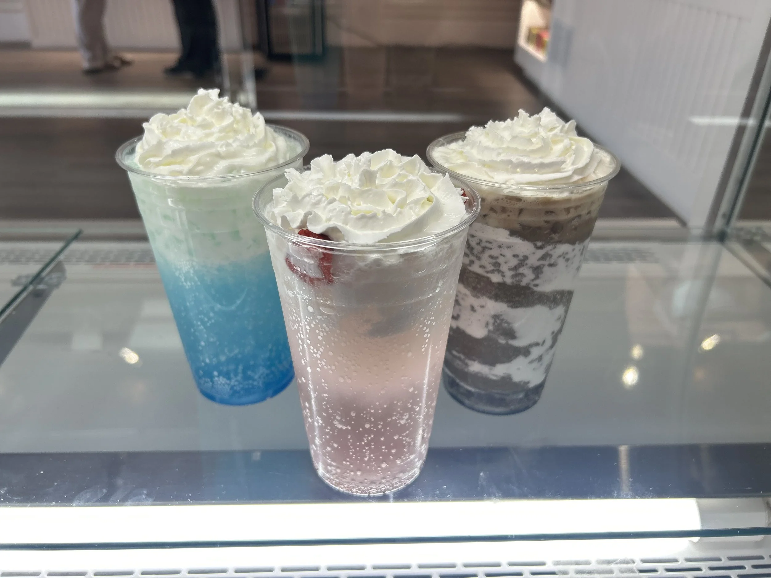 Three cups with whipped cream on top, containing colorful frozen drinks in shades of blue, pink, and brown.