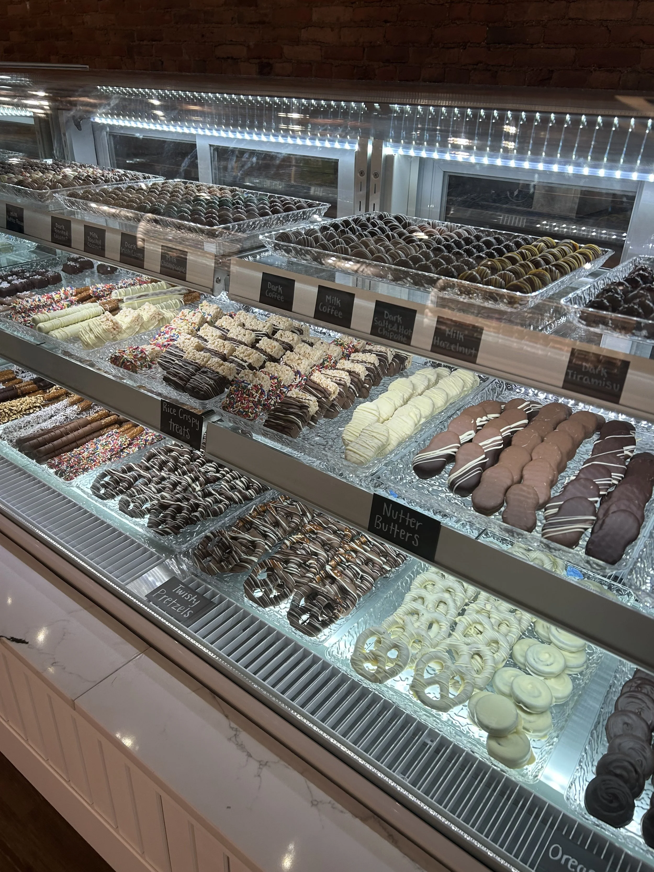 Display case filled with various types of decorated cookies, and chocolates, with labels for different flavors and toppings, such as dark chocolate, milk chocolate, and white chocolate.