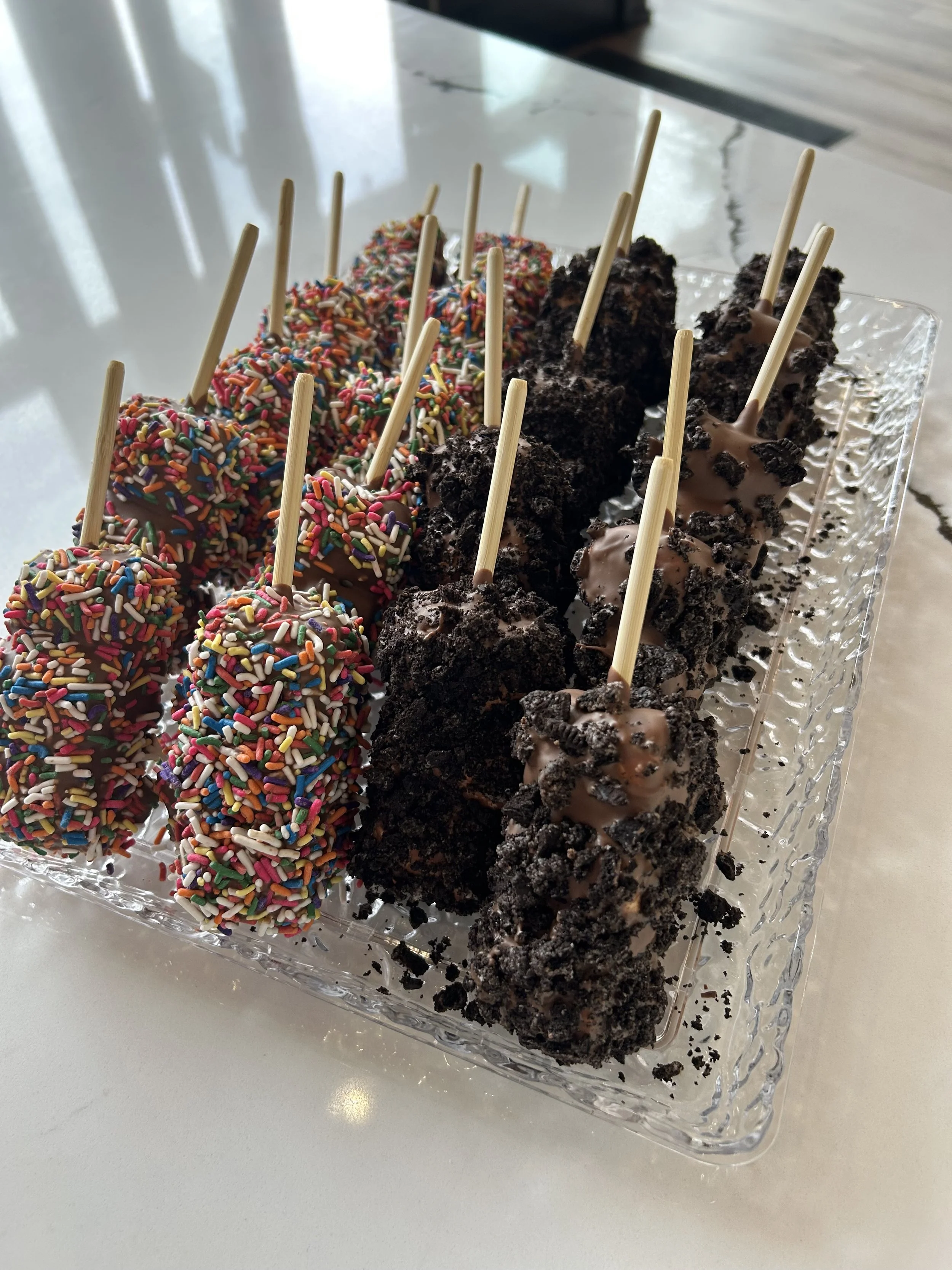 A glass platter with assorted chocolate covered marshmallows, some decorated with rainbow sprinkles and others coated with crushed cookies and chocolate, all with wooden sticks.