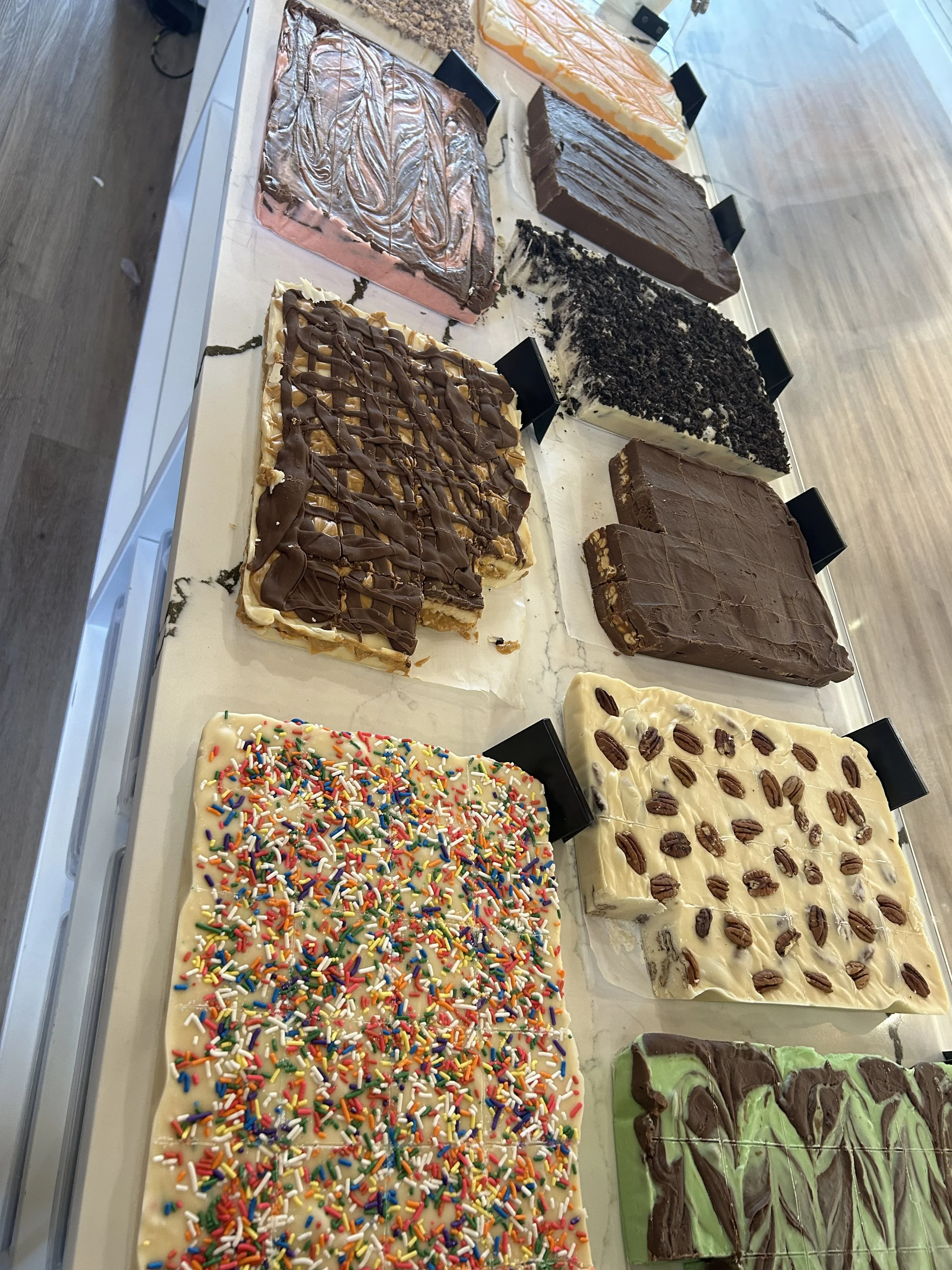 Assorted decorated slabs of fudge in a display case, featuring various flavors and toppings including sprinkles, chocolate, cookies, and fruit.