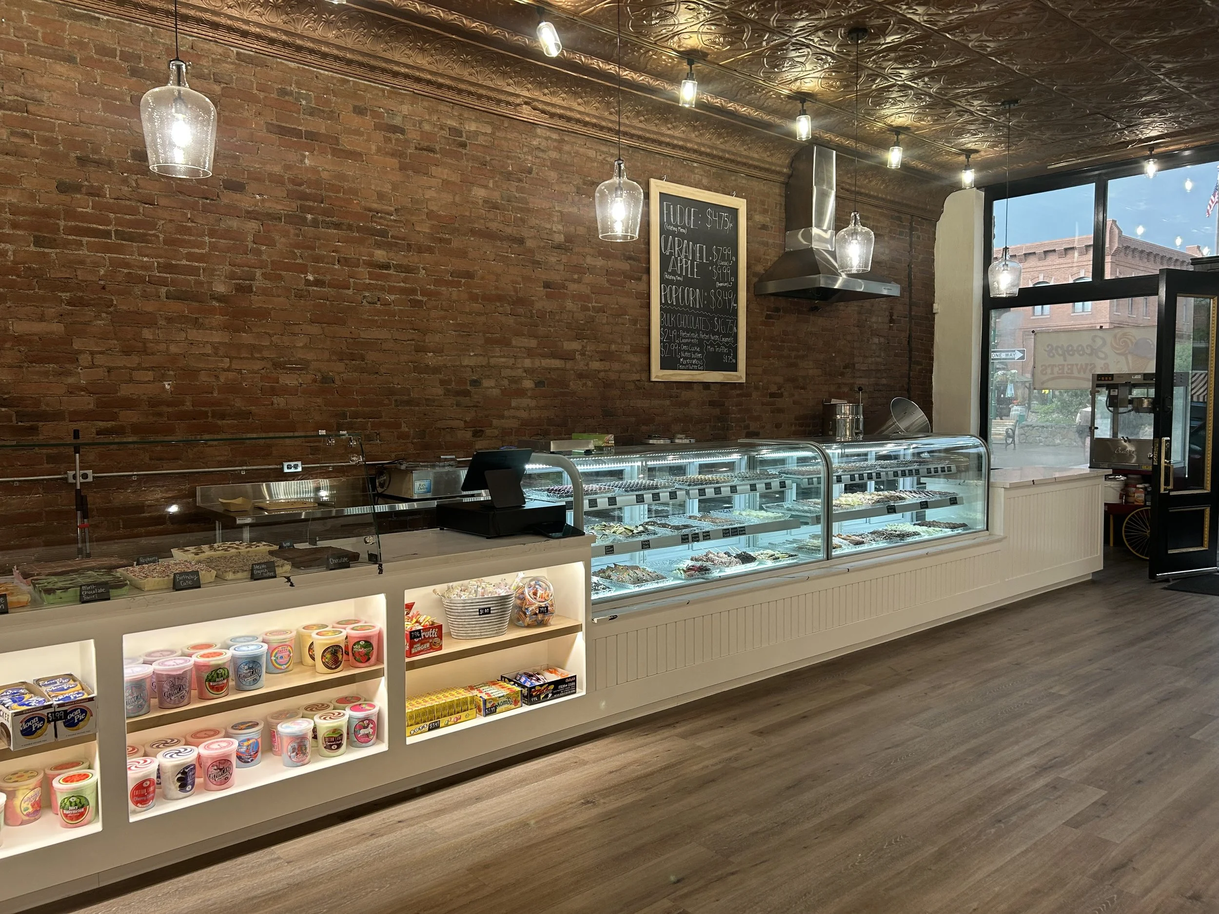 Interior of a candy shop featuring shelves of colorful candy jars, a glass display case with various sweets, a brick wall, a blackboard menu listing fudge, caramel apples, popcorn, and chocolate, and large windows showing a partial street view outsid