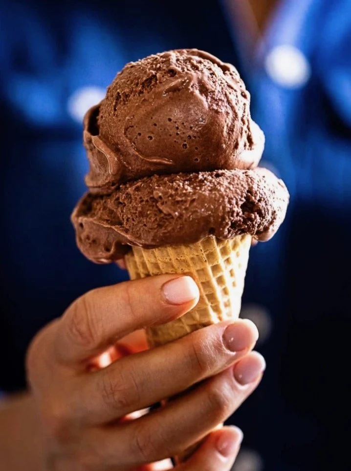 A person holding a cone with two scoops of chocolate ice cream.