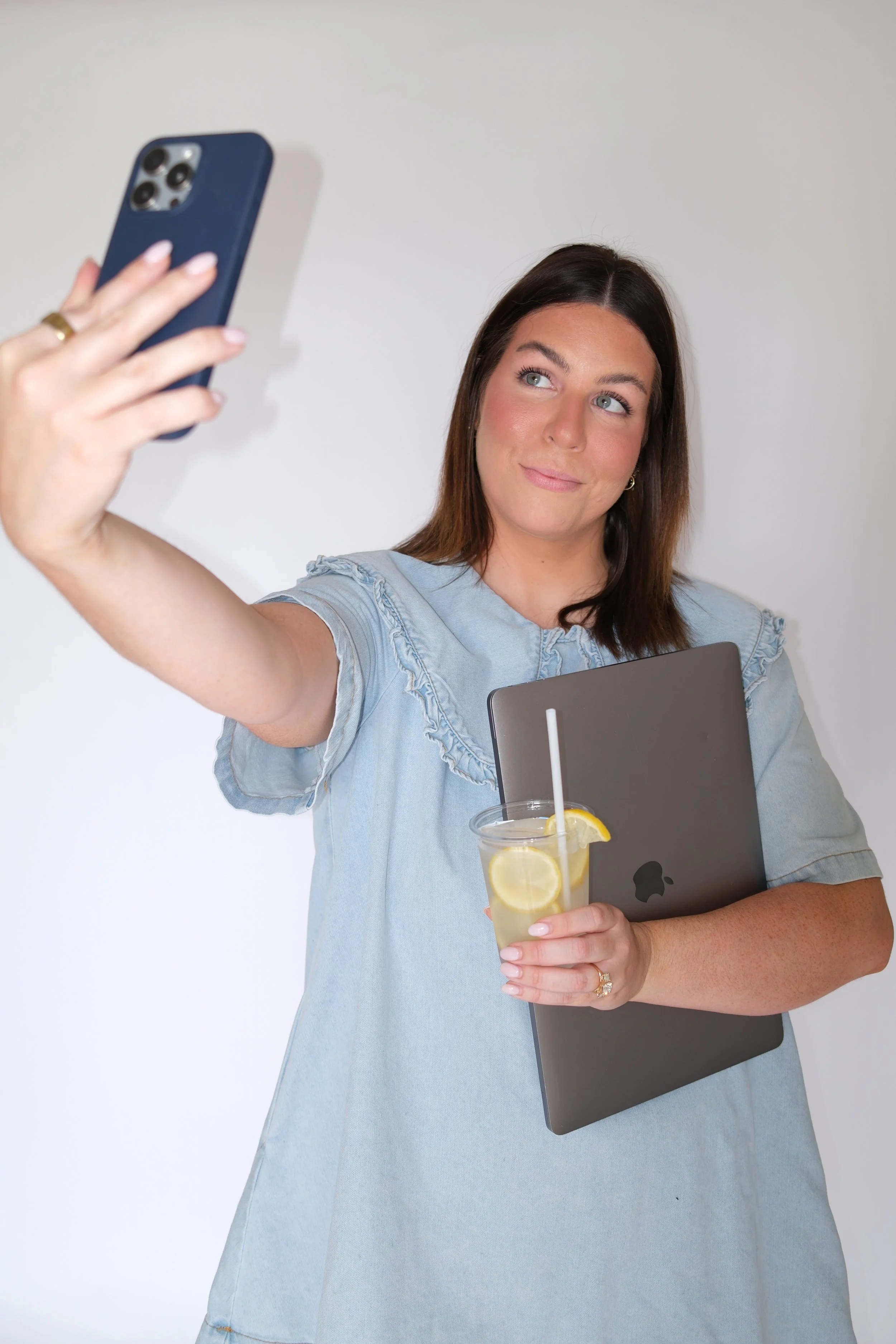 A woman taking a selfie with her phone, holding a laptop in one arm and a glass of lemon water in the other. She has brown hair, light skin, and is wearing a light blue shirt.