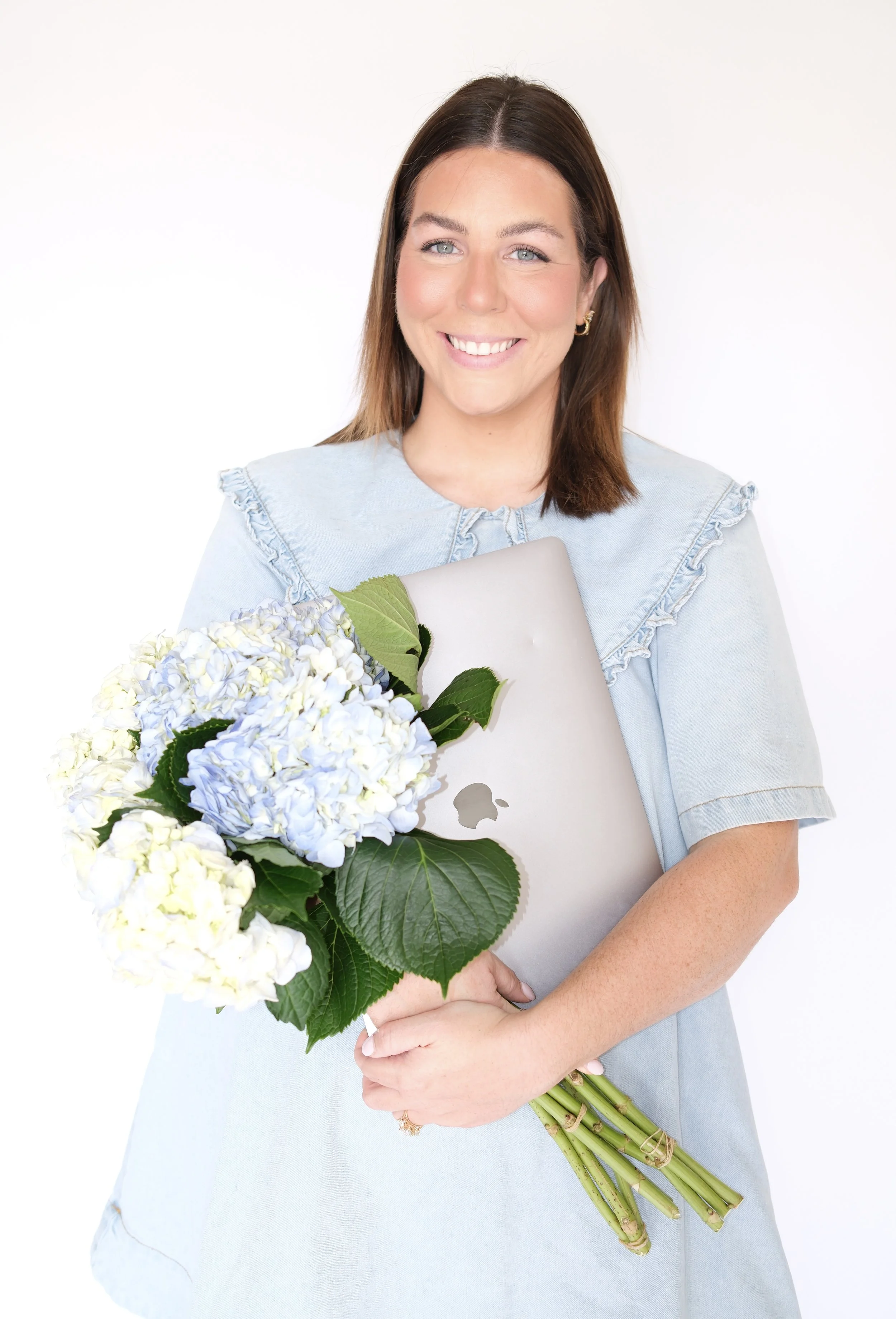 A woman smiling and holding a bouquet of white and light purple hydrangea flowers, along with a silver laptop.