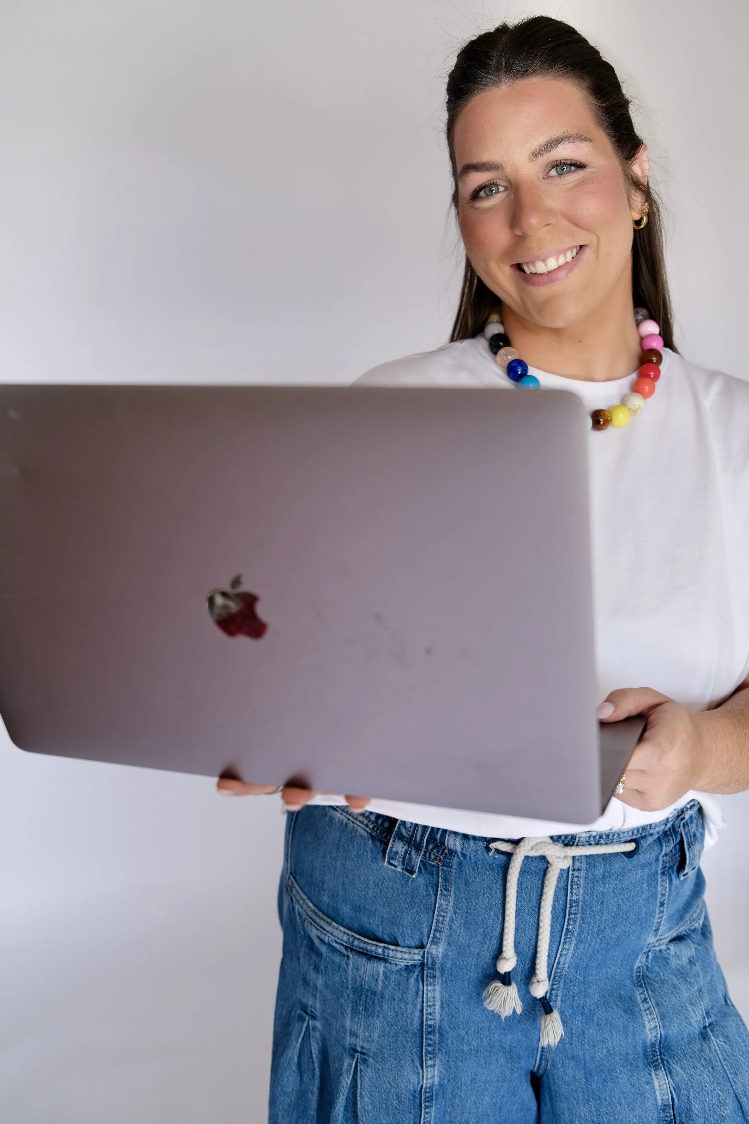 A woman with long brown hair, blue eyes, and a bright smile, holding a closed silver MacBook laptop. She is wearing a white T-shirt, blue jeans, and a colorful beaded necklace.