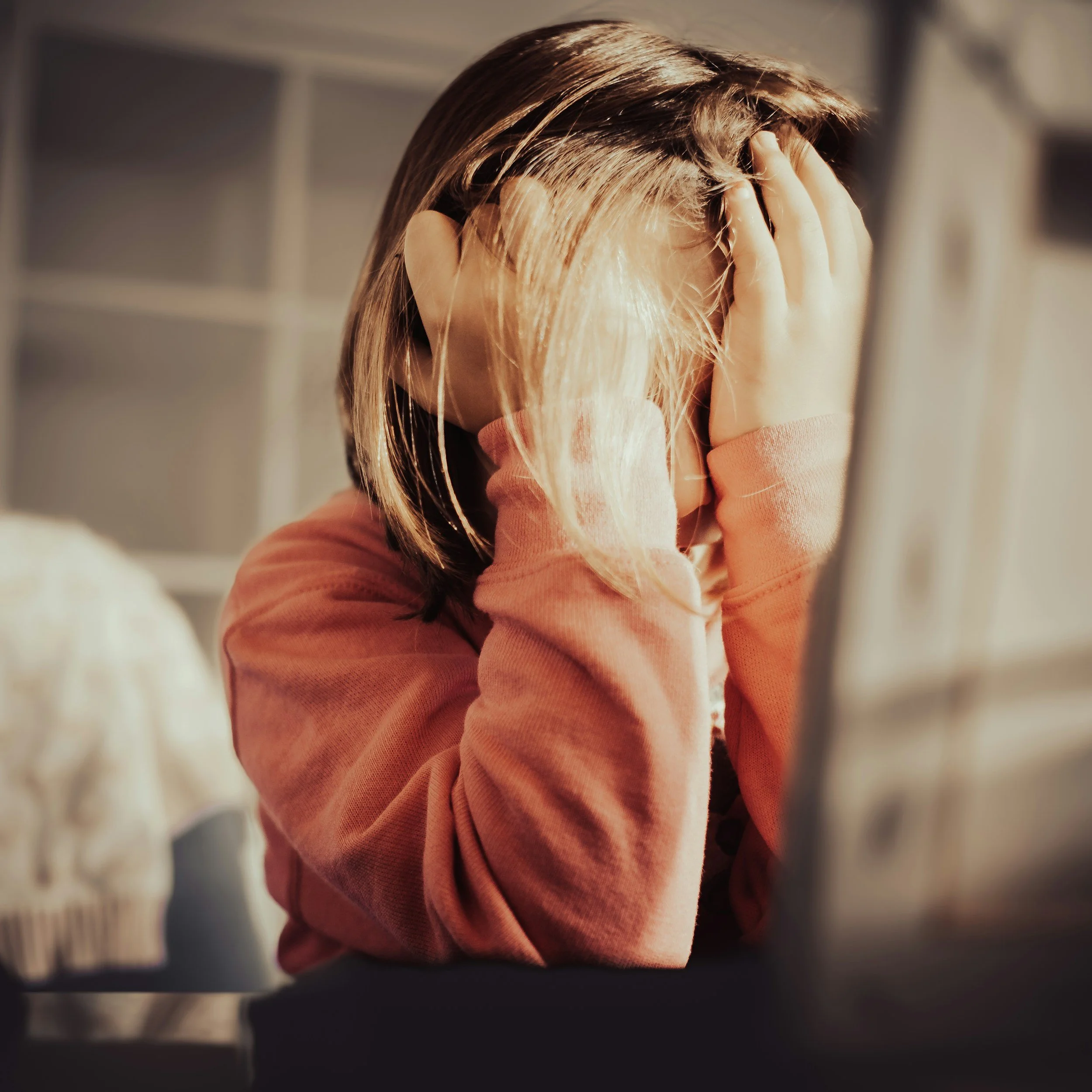 A person with long blonde hair holding their head with both hands, wearing a pink or peach sweater, with a blurred background.