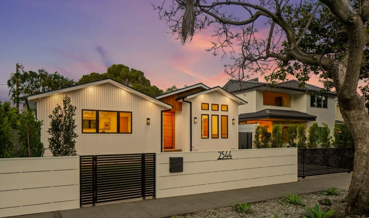 Modern house with white exterior walls, large windows, and a black fence at sunset.