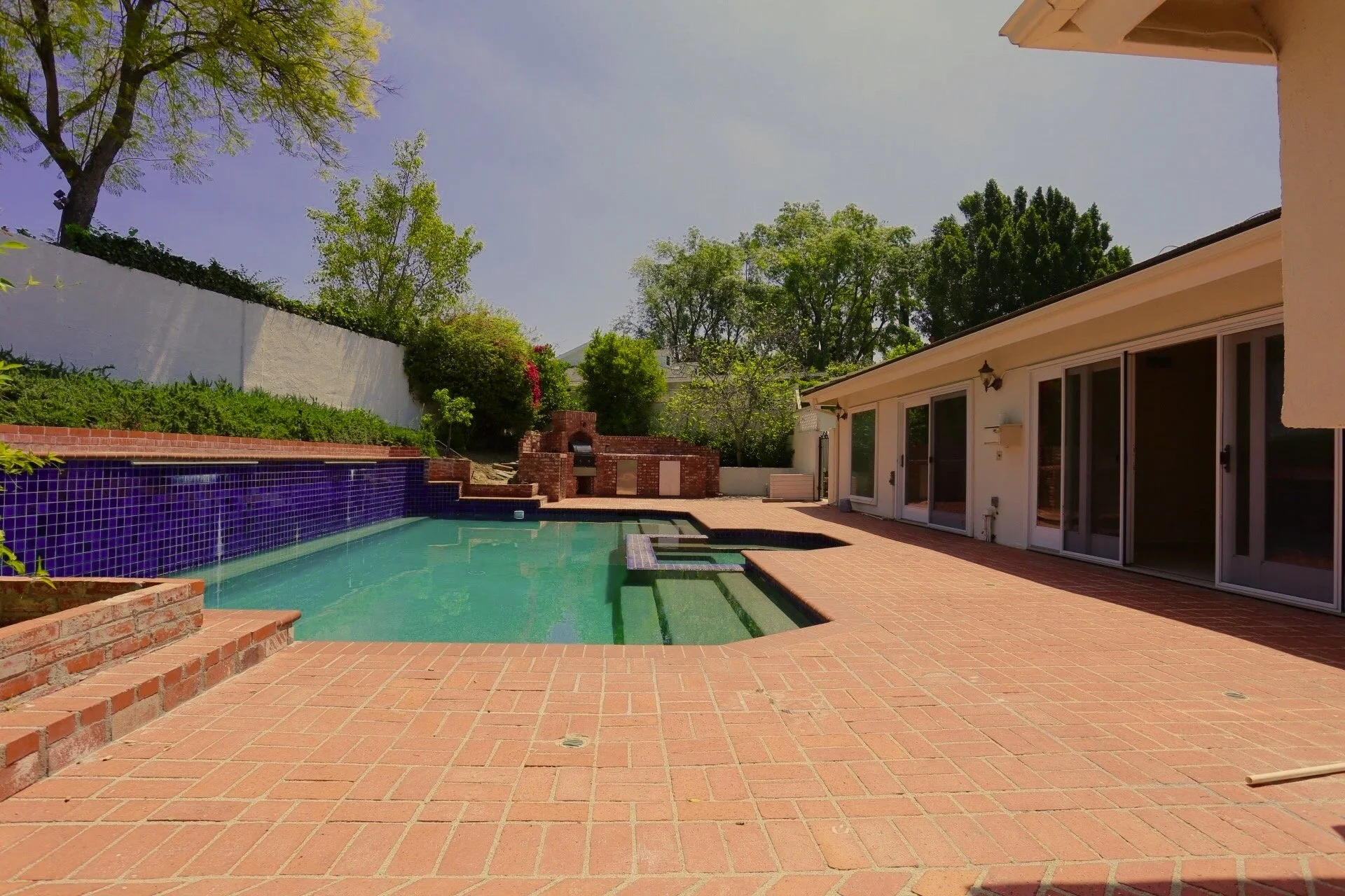Backyard with a swimming pool, brick patio, and a house with sliding glass doors. Green trees and a brick outdoor fireplace are visible in the background.