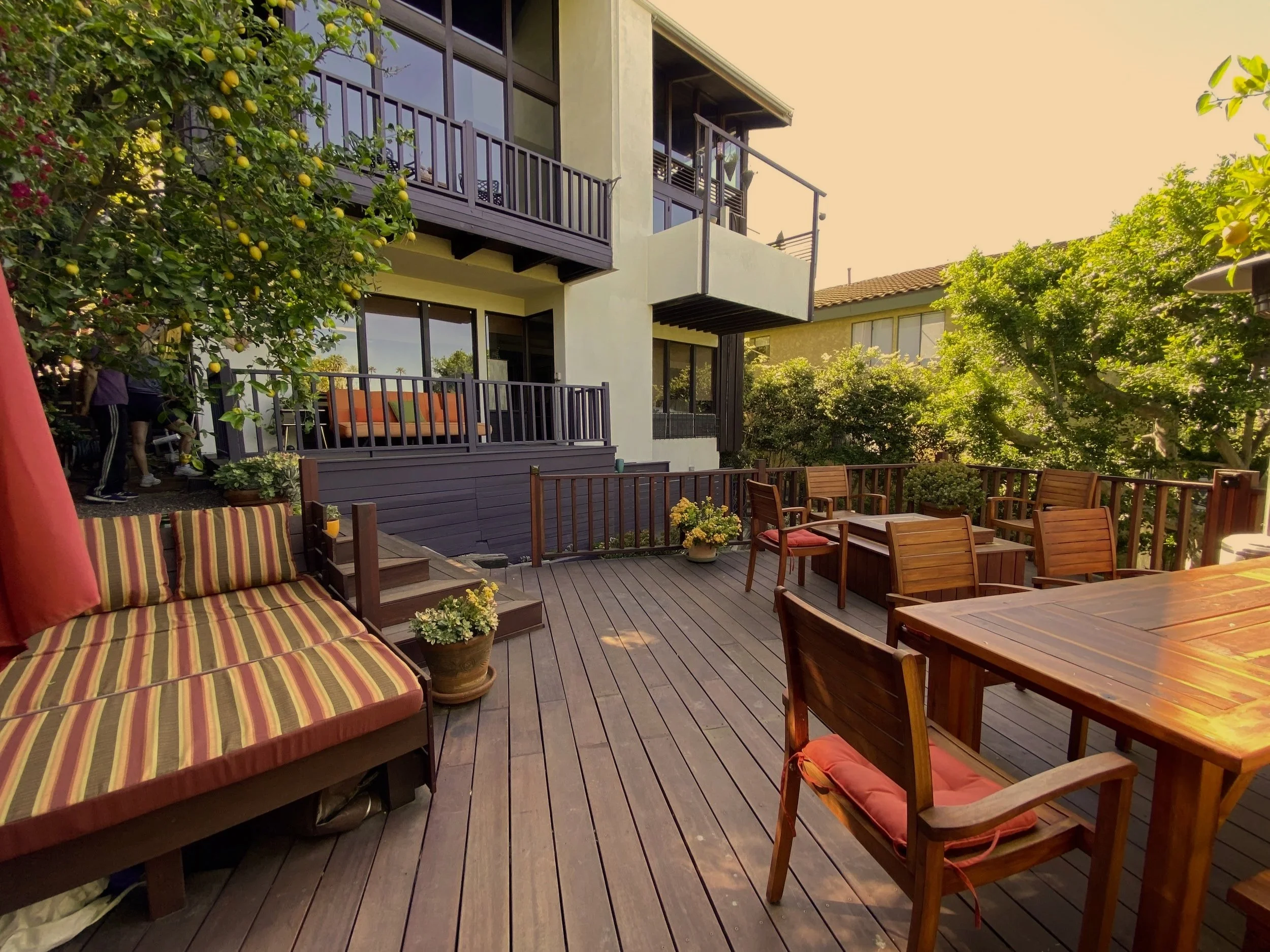 An outdoor patio area with wooden flooring, wooden table and chairs, and a lounge chair with striped cushions. There are potted plants, surrounding greenery, and a modern multi-story house with balconies and large windows in the background. The scene appears to be in the late afternoon or early evening, with warm sunlight.