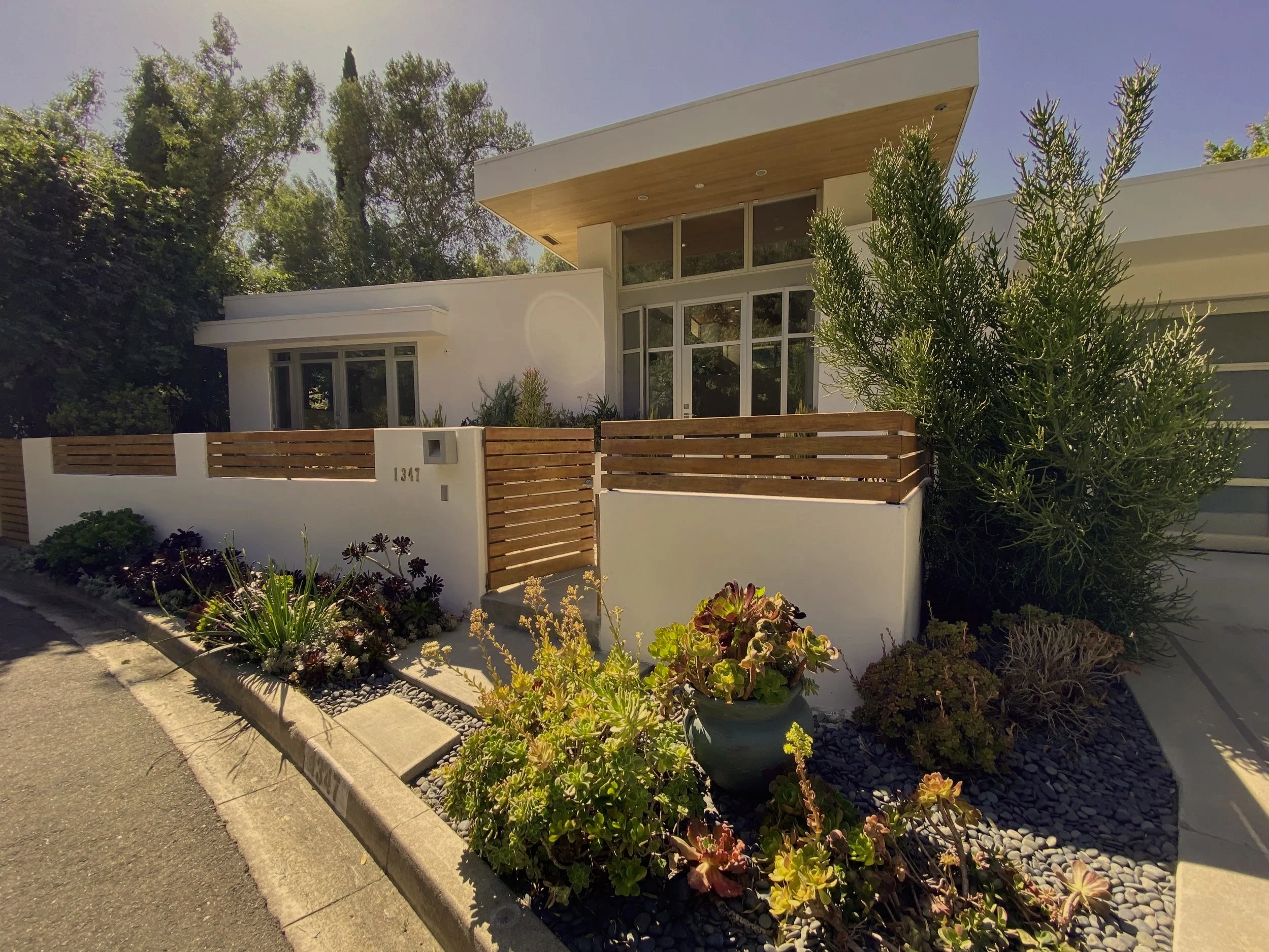Modern white house with large glass windows, surrounded by lush green trees and a garden with various plants and flowers, in sunlight.
