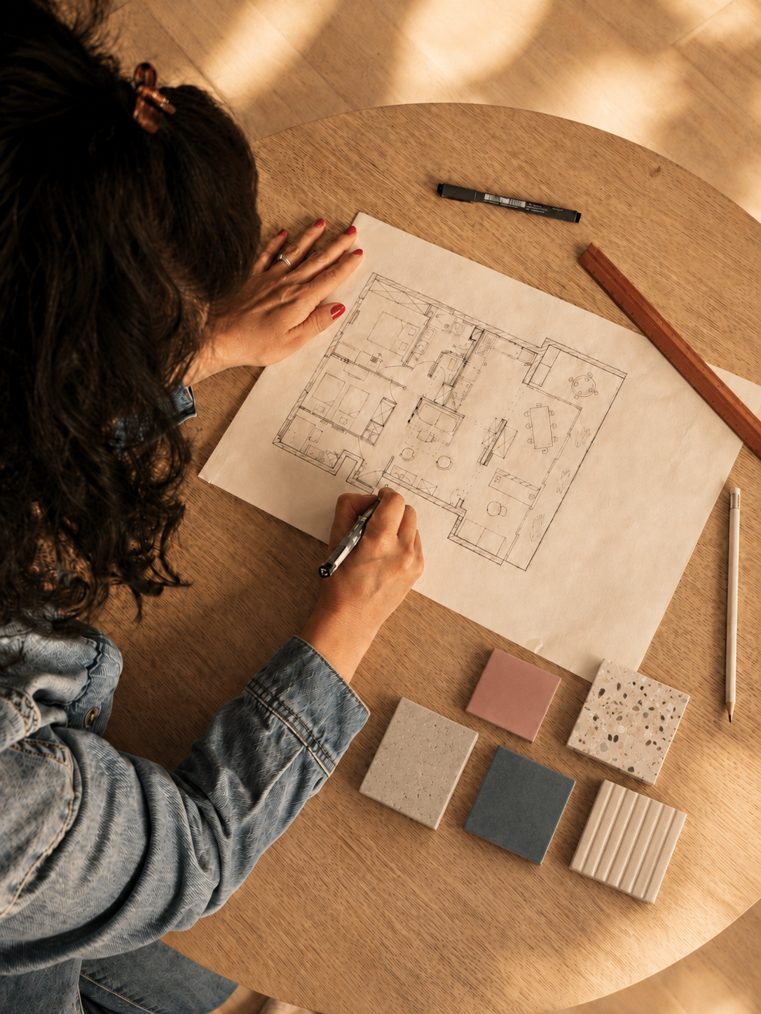 Une personne dessinant un plan d'architecture sur une table en bois, avec des échantillons de matériaux et des outils.