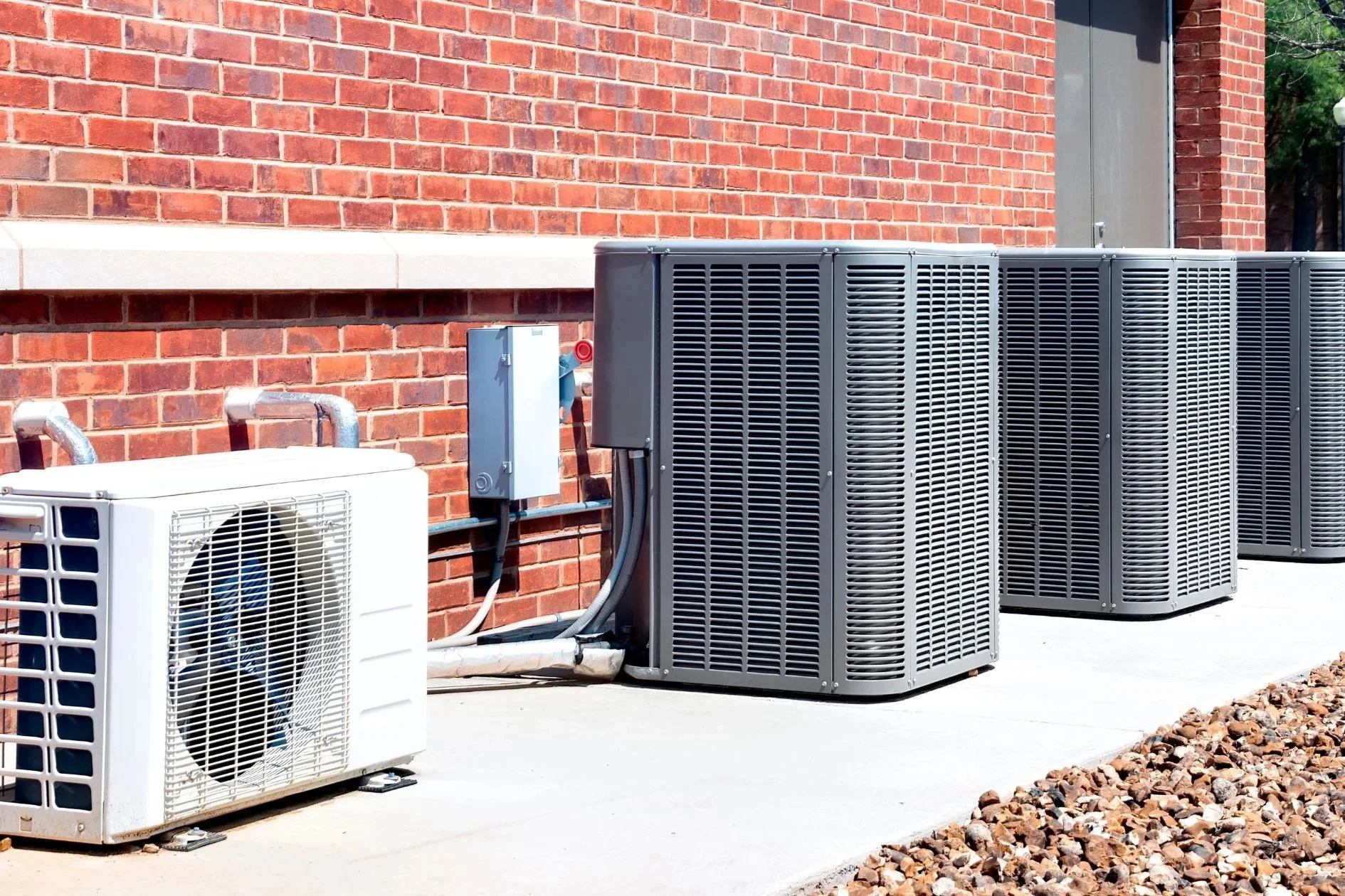 Outdoor scene with multiple HVAC units placed on a concrete surface against a red brick wall, with some gravel in the foreground.