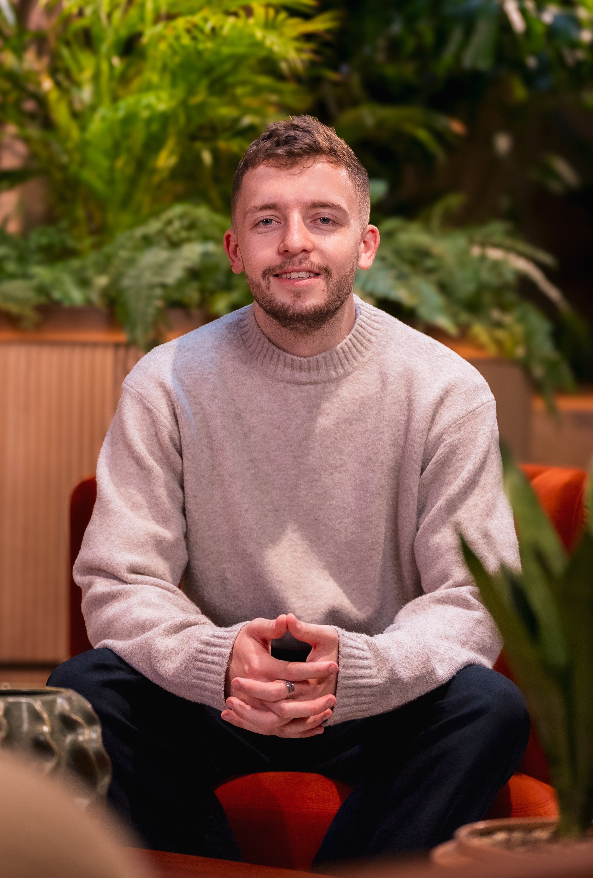 A young man with light brown hair, beard, and mustache, wearing a beige knitted sweater, sitting in a cozy indoor setting with green plants in the background, smiling softly, and clasping his hands in his lap.