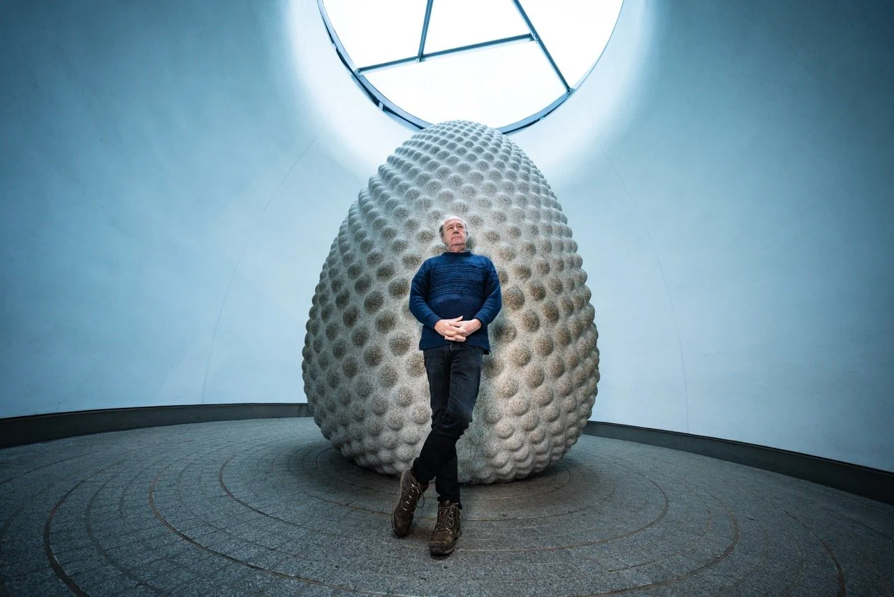 A man standing in front of a large, textured sculpture resembling a golf ball, inside a modern, circular room with a curved wall and a skylight overhead.