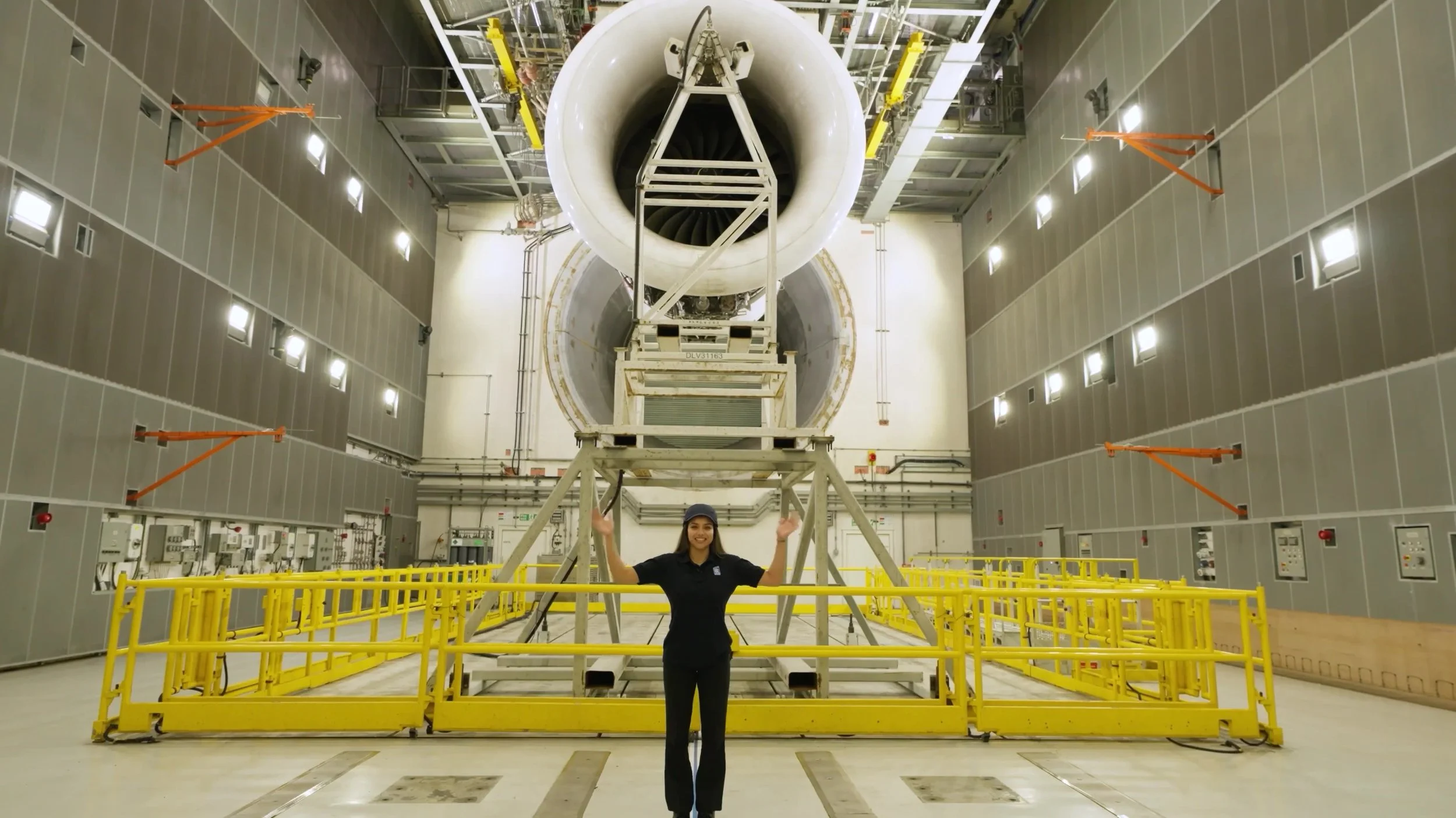 A woman is standing in front of a large jet engine inside an industrial facility, giving a thumbs up.