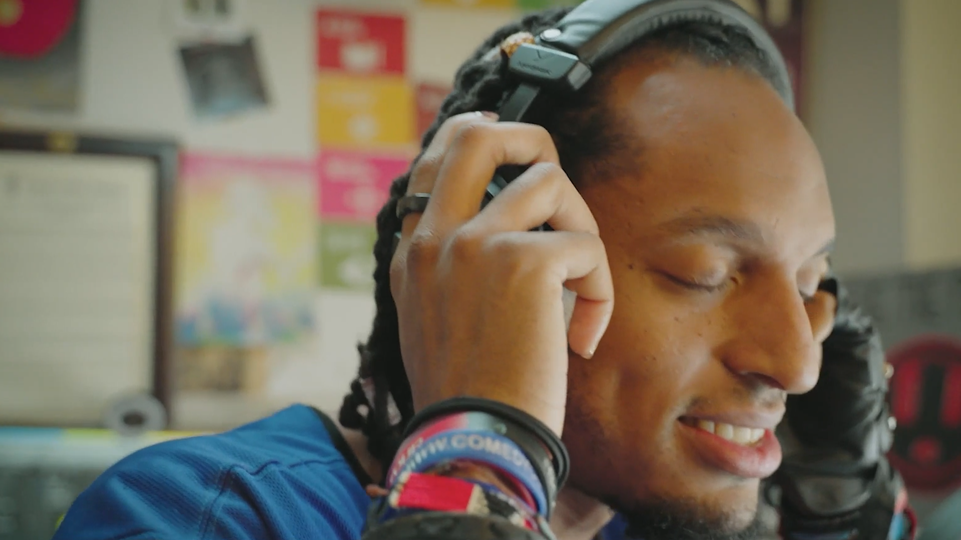 A person wearing headphones, smiling with eyes closed, and wearing a blue jacket, in a classroom with colorful posters and books in the background.