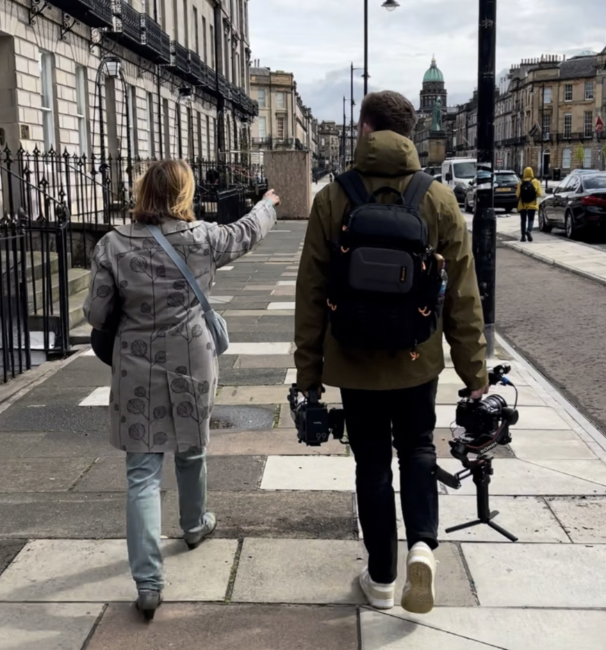 Two people walking on a city sidewalk, with one person pointing ahead. One person is carrying a camera and a drone, both in their hands. The street is lined with historic buildings, parked cars, and a domed structure in the background.