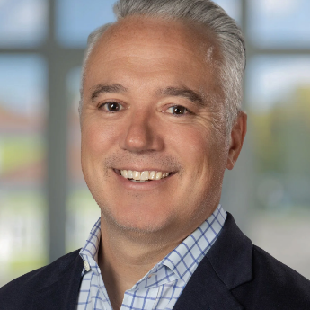 A smiling middle-aged man with gray hair, wearing a checkered shirt and a dark blazer, standing indoors with a bright window in the background.