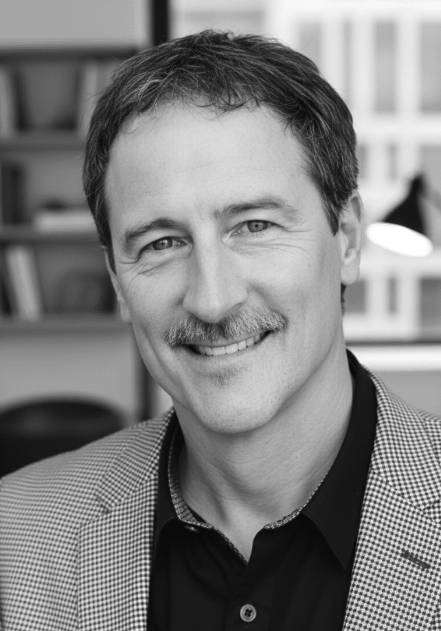 Black and white portrait of a smiling man with short hair, a mustache, wearing a checkered blazer and black shirt, background blurred with bookshelves.
