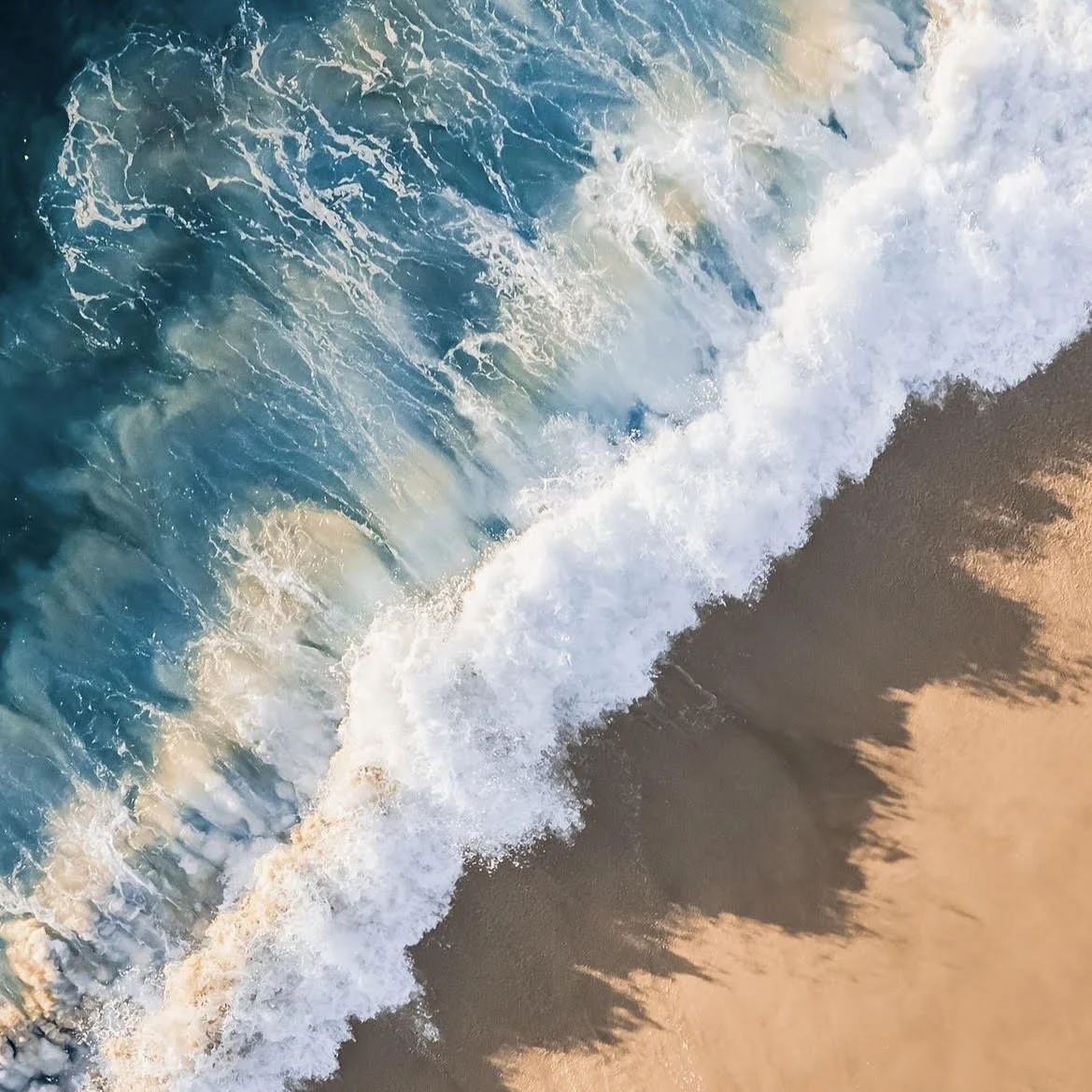 Aerial view of ocean waves crashing onto sandy beach at sunset, with foamy white surf and shadow of a person or object on the sand.