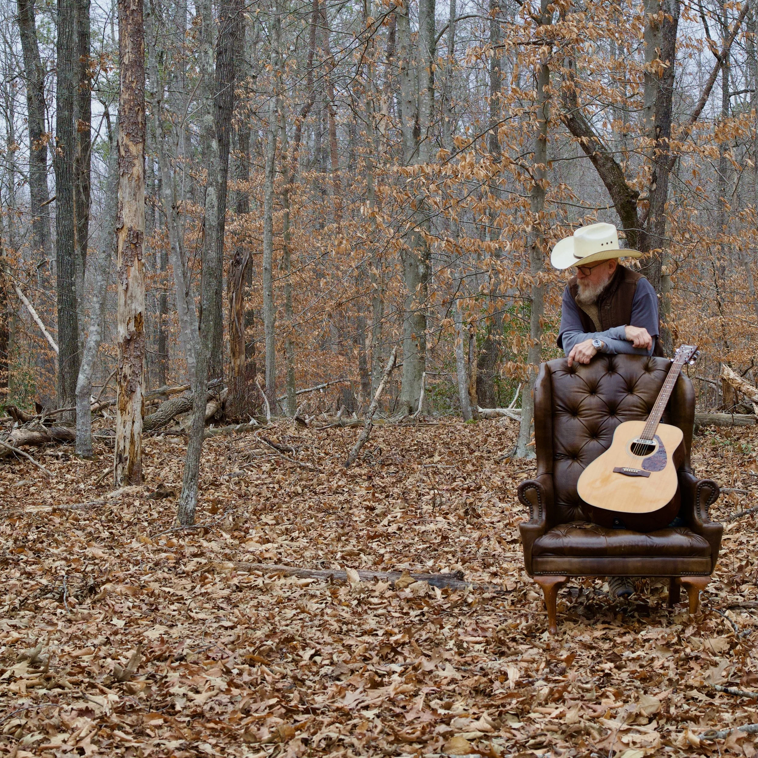 Man with a white cowboy hat and glasses, sitting on a brown leather tufted armchair in a woodland setting with dry leaves on the ground and an acoustic guitar.
