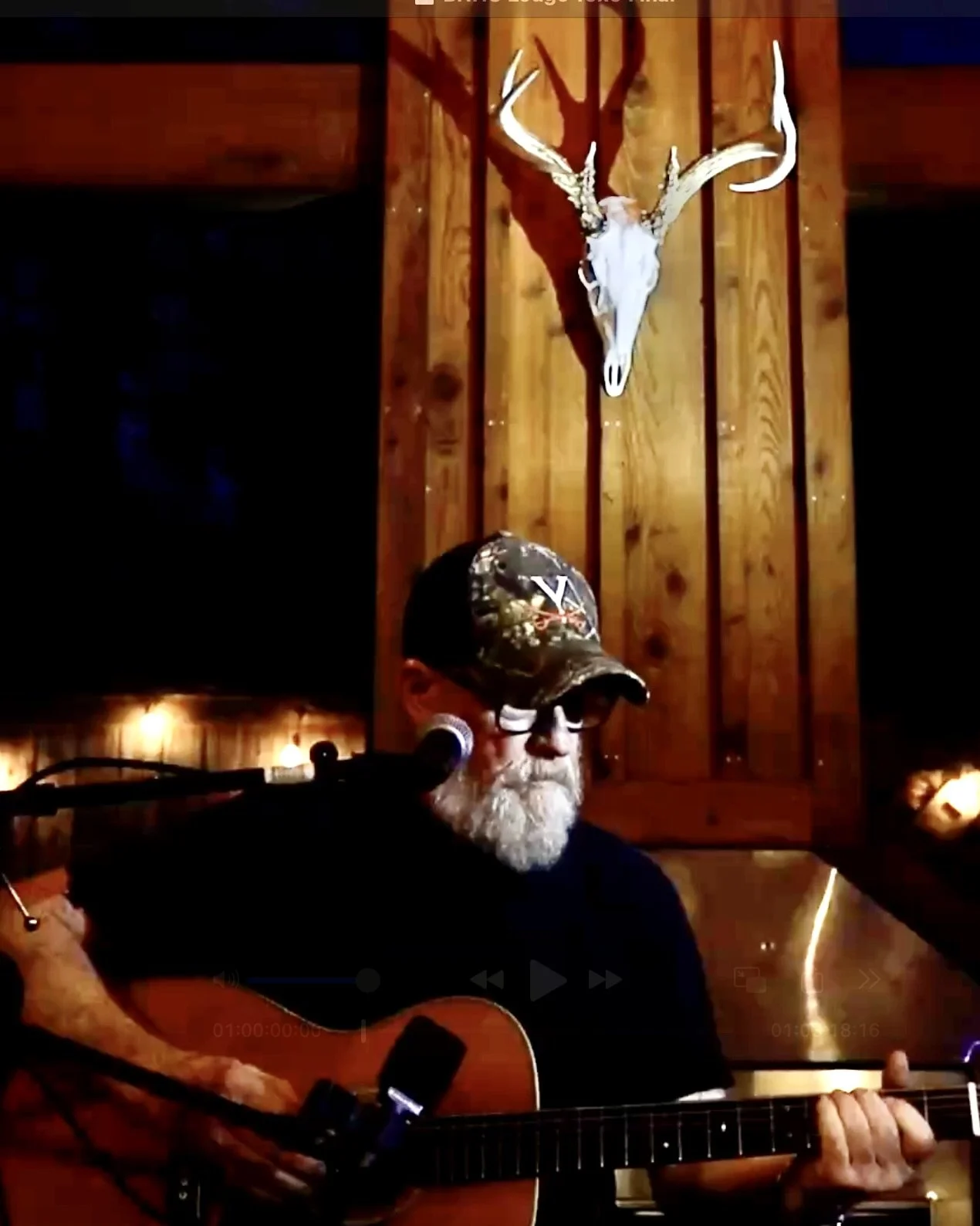 A man with a white beard wearing a camouflage baseball cap and sunglasses, playing an acoustic guitar and singing into a microphone, in a rustic wooden room with a deer skull with antlers mounted on the wall behind him.