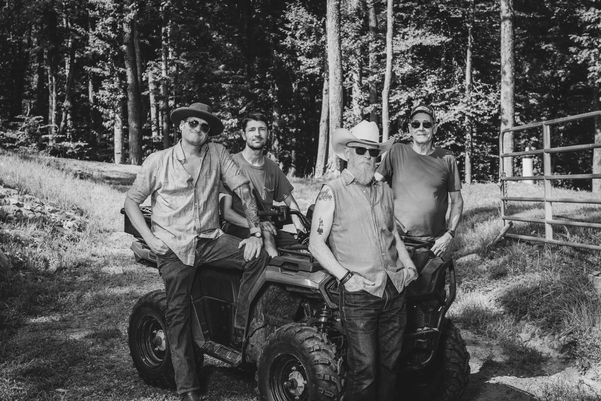 Four men standing outdoors in a forested area, with one on an all-terrain vehicle and the others standing beside it, all wearing sunglasses and casual clothing, some with hats.