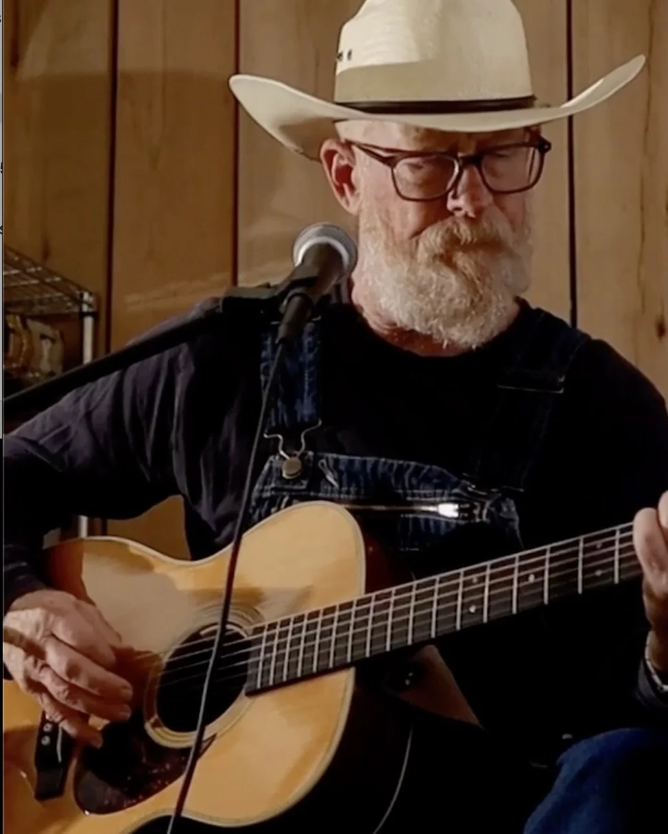 An older man with glasses and a cowboy hat playing an acoustic guitar in front of a wooden wall.
