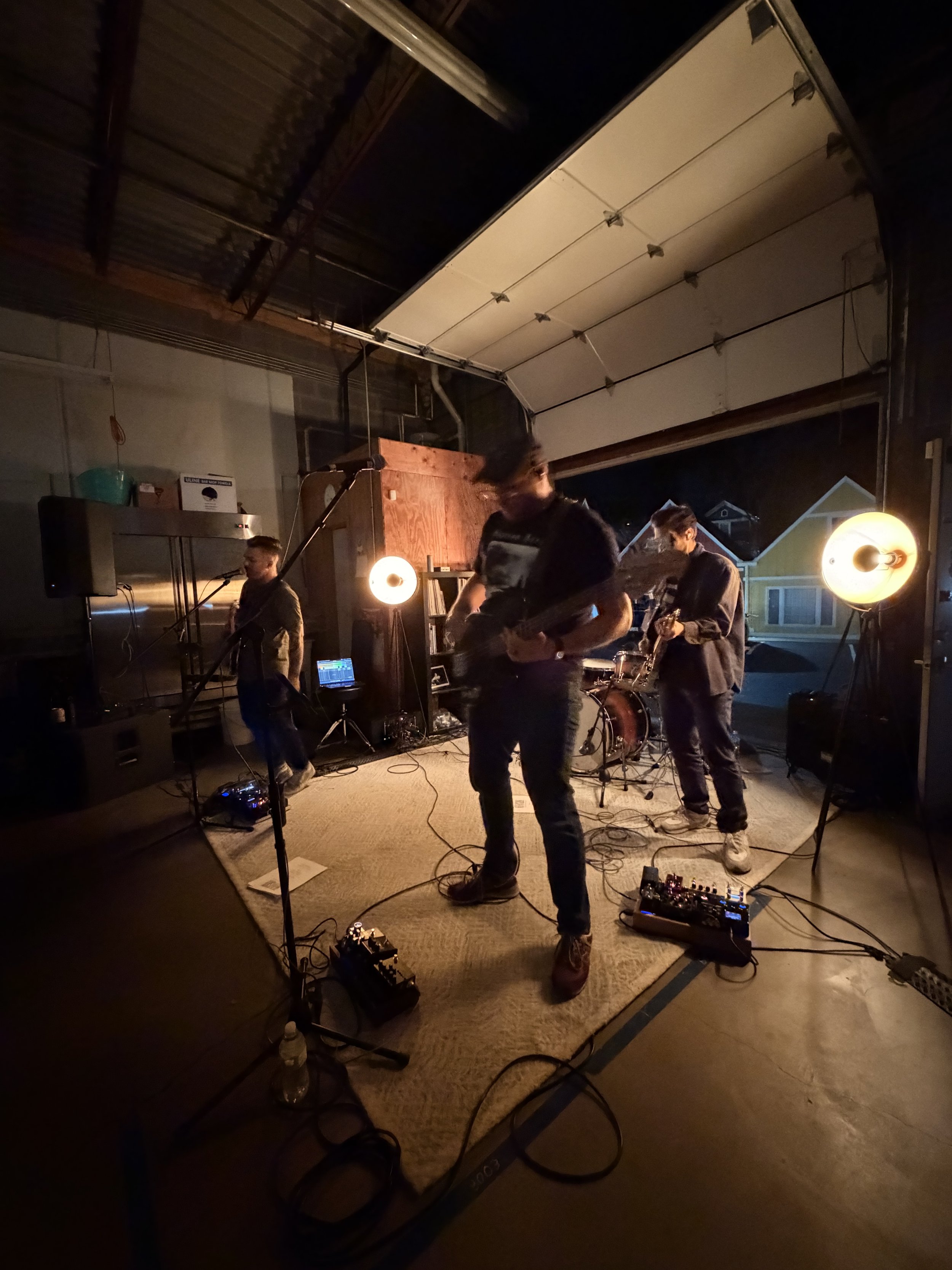 A band performing in a garage at night, with three musicians playing instruments and various sound equipment set up around them.