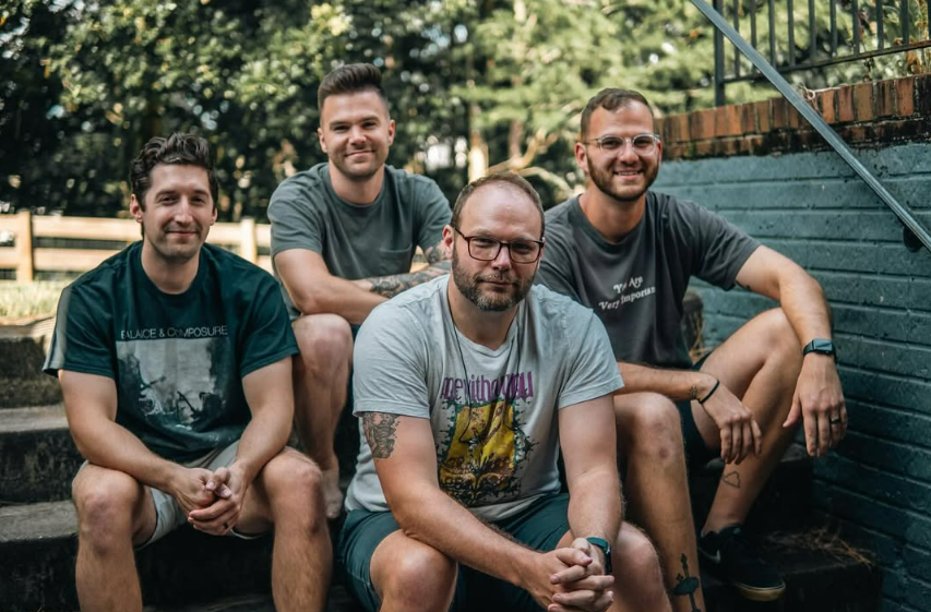 Four men sitting on outdoor stairs in a backyard with trees and a fence in the background.