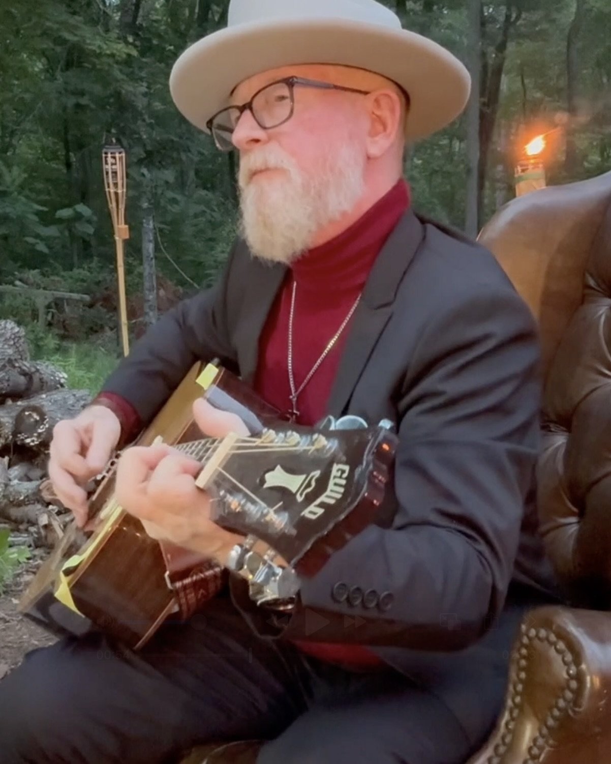 An older man with a white beard, glasses, and a wide-brimmed hat playing an acoustic guitar outdoors in a wooded area, seated on a leather chair.