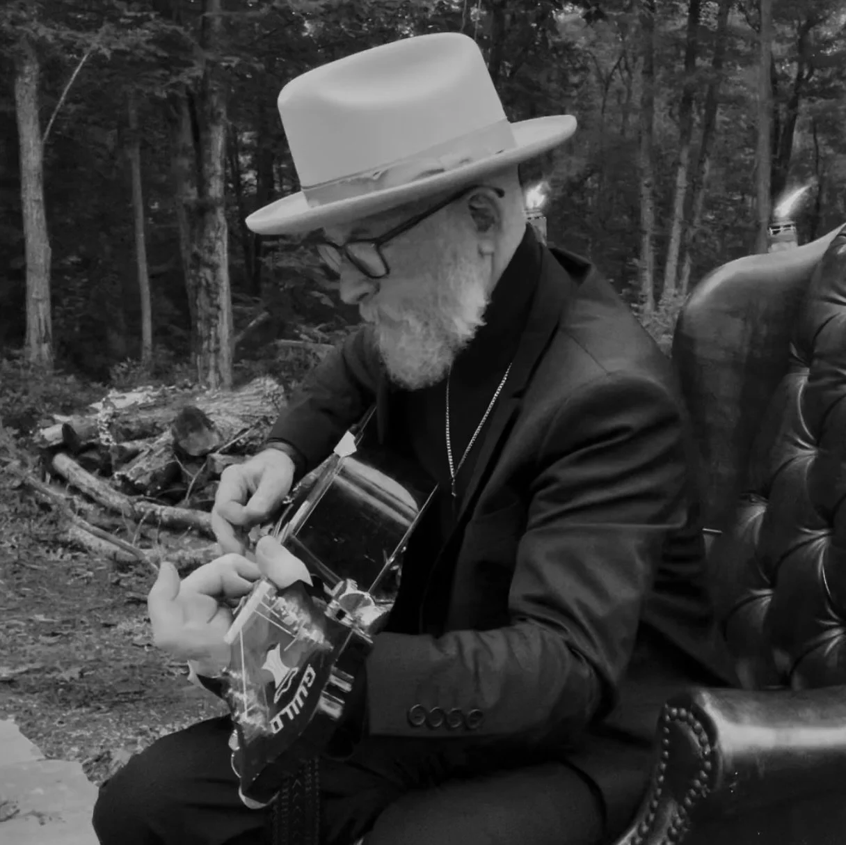 An elderly man with a white beard and glasses, wearing a wide-brimmed hat and a black blazer, is sitting outdoors in a forested area. He is holding a guitar and a small plastic bag, looking down at the guitar while playing or tuning it.