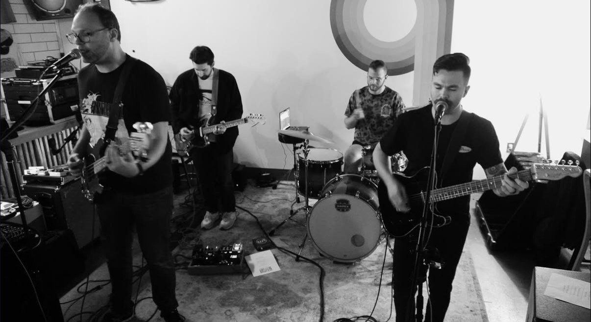 Four musicians performing with guitars and drums in a room.