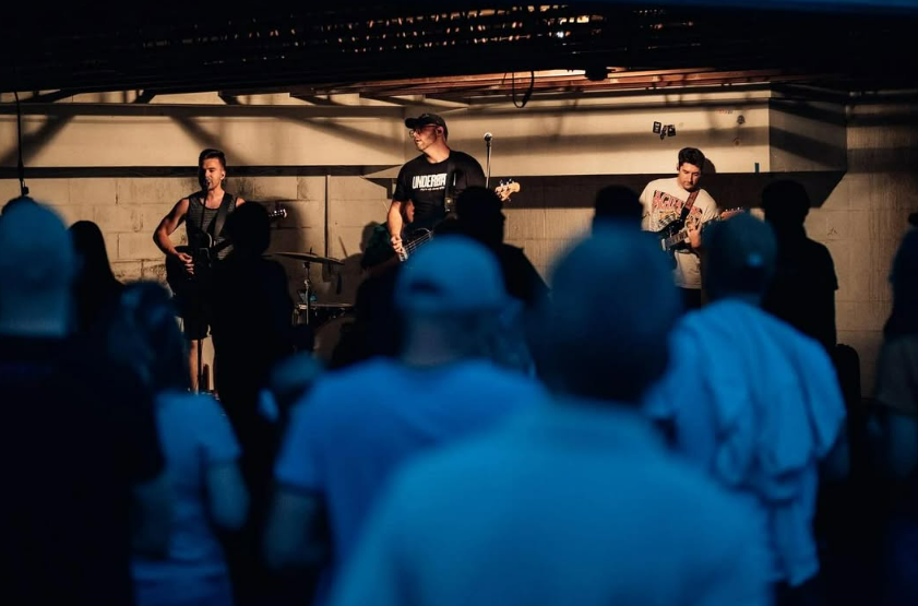 A band performs on stage at a concert venue, with three members playing guitars and one playing drums, while the audience watches.